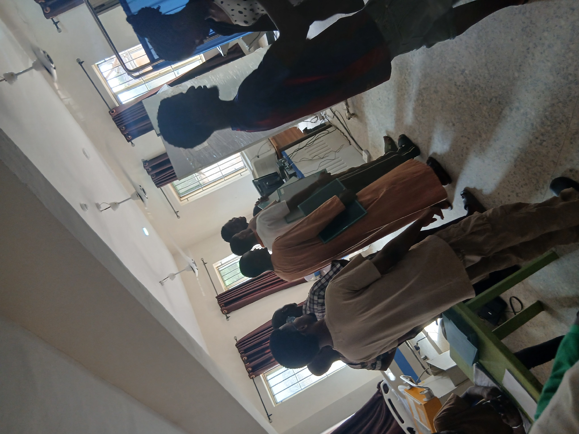 The image shows a classroom in Lagos, Nigeria. The room is brightly lit by daylight coming through several windows. There are people sitting at tables, likely students, with books or tablets in front of them. Some are wearing uniforms and are arranged in rows. The room has ceiling fans.