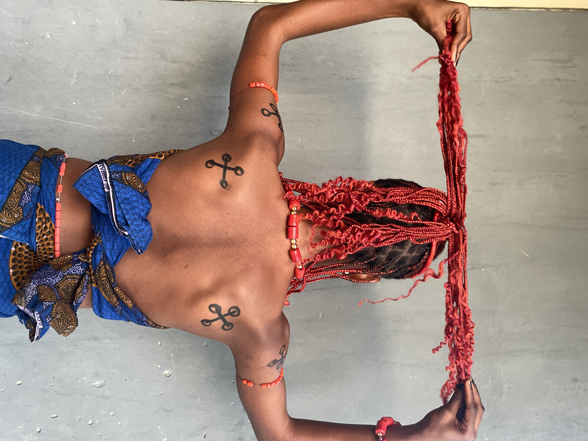 A high-angle full shot depicts an individual, presumably female, wearing red-orange braided hair extensions. The person is adorned with red-orange beaded jewelry and what appears to be body paint, featuring a cross design, and a blue garment. The person stands or kneels on a concrete surface indoors. The location may be in Badagry, Nigeria. The individual is holding the extensions up with both hands. The person's back is partially exposed.