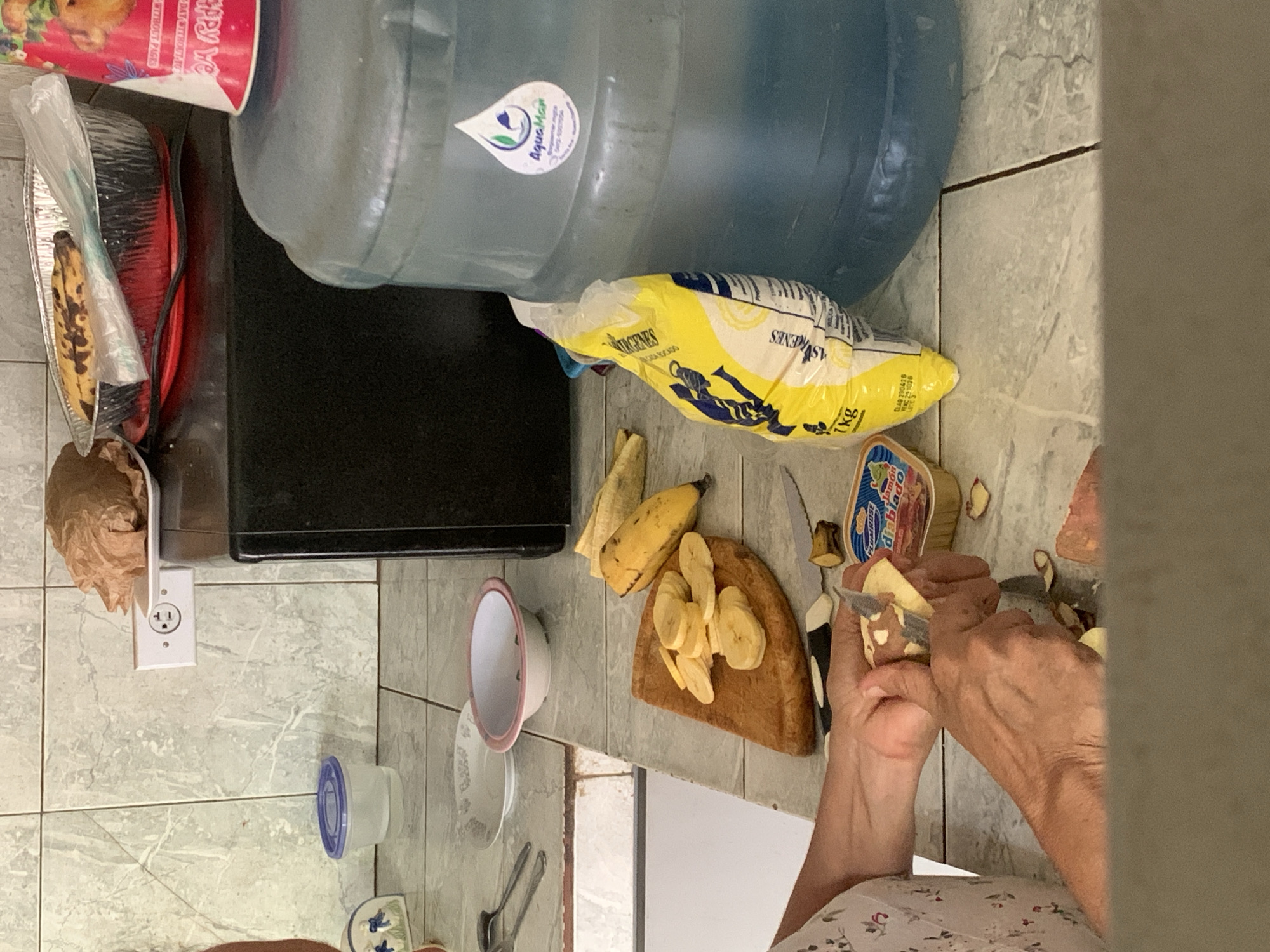 An overhead view captures a person's hands in the process of peeling a banana on a kitchen counter. The hands, appearing to belong to an older individual, use a small knife to remove the peel from a banana. To the left, a wooden cutting board holds several round slices of banana and another whole, partially peeled banana, alongside a knife with a black and white handle.
The kitchen counter is a light-colored, possibly tiled, surface. Various household items are scattered around. A white bowl with a pink rim and a clear plastic container with a blue lid are visible to the left of the cutting board. Further back and left, a white electrical outlet is mounted on the wall.
In the upper part of the image, a large, translucent blue water bottle is prominent, featuring an "AquaMan" label with the number "SAS-5007504". Adjacent to it is a yellow bag, likely containing flour or a food mix, with "1kg" and fragmented text such as "ASVRENES URGENES CICA KACADO" visible. A red and white container, possibly a food package, displays partial text including "appy Ye" and "WITHOUT PAGES". Resting in a silver foil tray is a banana with dark spots. A small jar labeled "Jamon Diablo" (deviled ham) from the "Plumrose" brand is also visible near the hands. The floor is tiled with light grey, patterned tiles, indicating a home kitchen environment. The visible brands and items suggest a location in Latin America, consistent with Santa Ana, Venezuela.