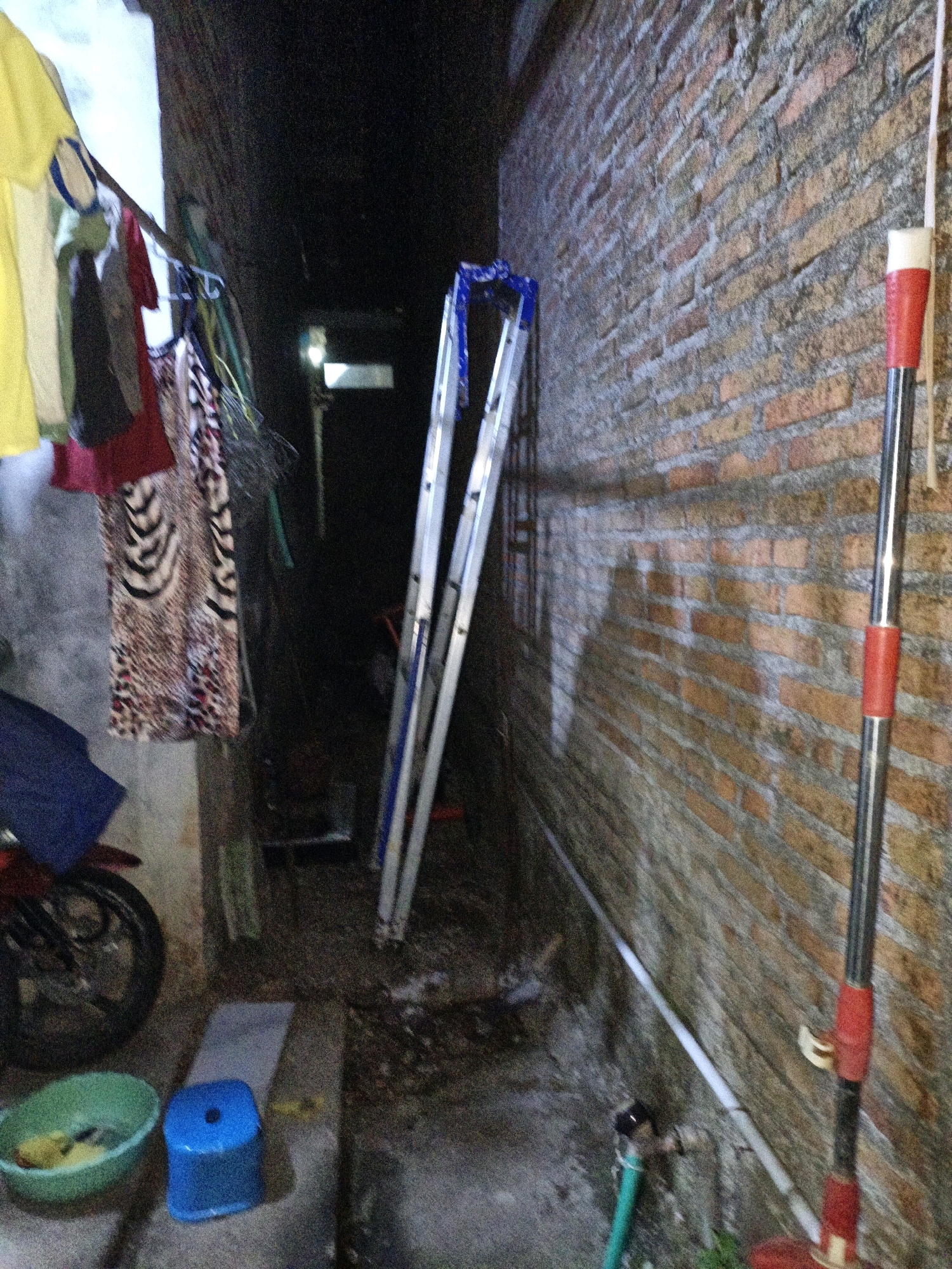 The image depicts a dimly lit outdoor alleyway. On the left, laundry hangs on a clothesline, including a yellow shirt, a patterned dress, and other garments. A blue plastic stool sits on the ground, next to a green plastic basin containing what appears to be yellow fabric. In the background, a ladder is propped against a brick wall. To the right, a broom with a red handle leans against the same wall. The ground is a mix of concrete and dirt. The overall scene suggests a residential area. The location is identified as Cikal, Indonesia.