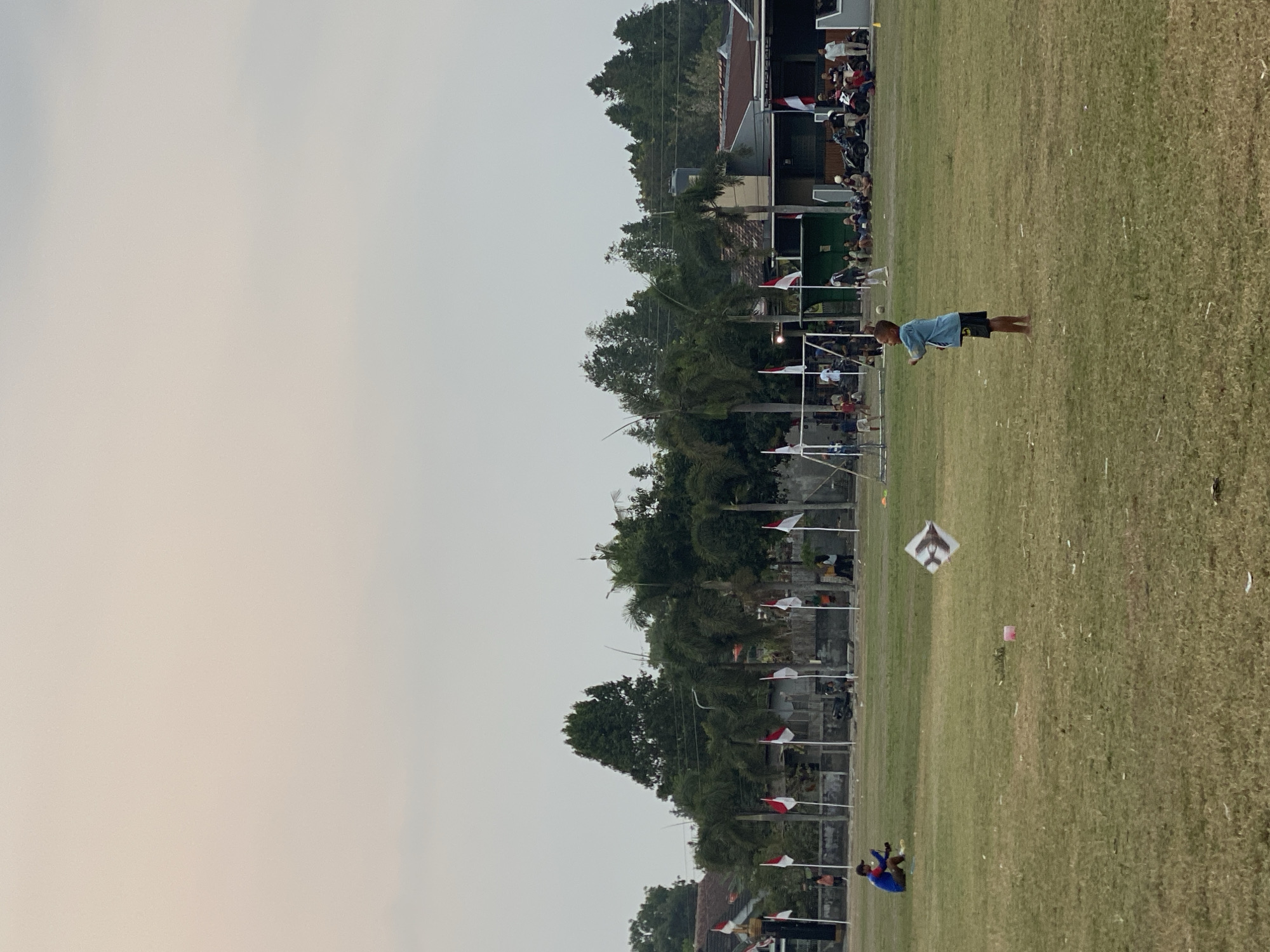 The image depicts an outdoor scene in the city of Kasihan, Indonesia, likely on a sports field or large open grassy area. The time of day appears to be late afternoon or early evening, given the soft, diffused lighting and the muted colors of the sky.
In the foreground, a young boy is standing on the grass, looking to his right with his arms slightly raised, as if he is about to throw something or has just thrown it. He is barefoot and wearing a light blue t-shirt and dark shorts. A kite, shaped like an airplane, is visible in the air to his left, suggesting he might be involved in flying it.
Further in the background, a man is seated on the grass to the left of the frame, appearing to be holding a kite string. He is wearing a blue and red shirt and dark pants. The field itself is covered in dry, greenish-yellow grass with some scattered debris.
Along the far edge of the field, a line of Indonesian flags (red and white) on poles marks a boundary. Behind the flags, there are buildings and trees. Some people are visible in the distance, gathered near the flags and in front of the buildings. There are also some parked motorcycles and what appears to be a soccer goal structure. The overall atmosphere is relaxed and appears to be a community gathering or recreational event.