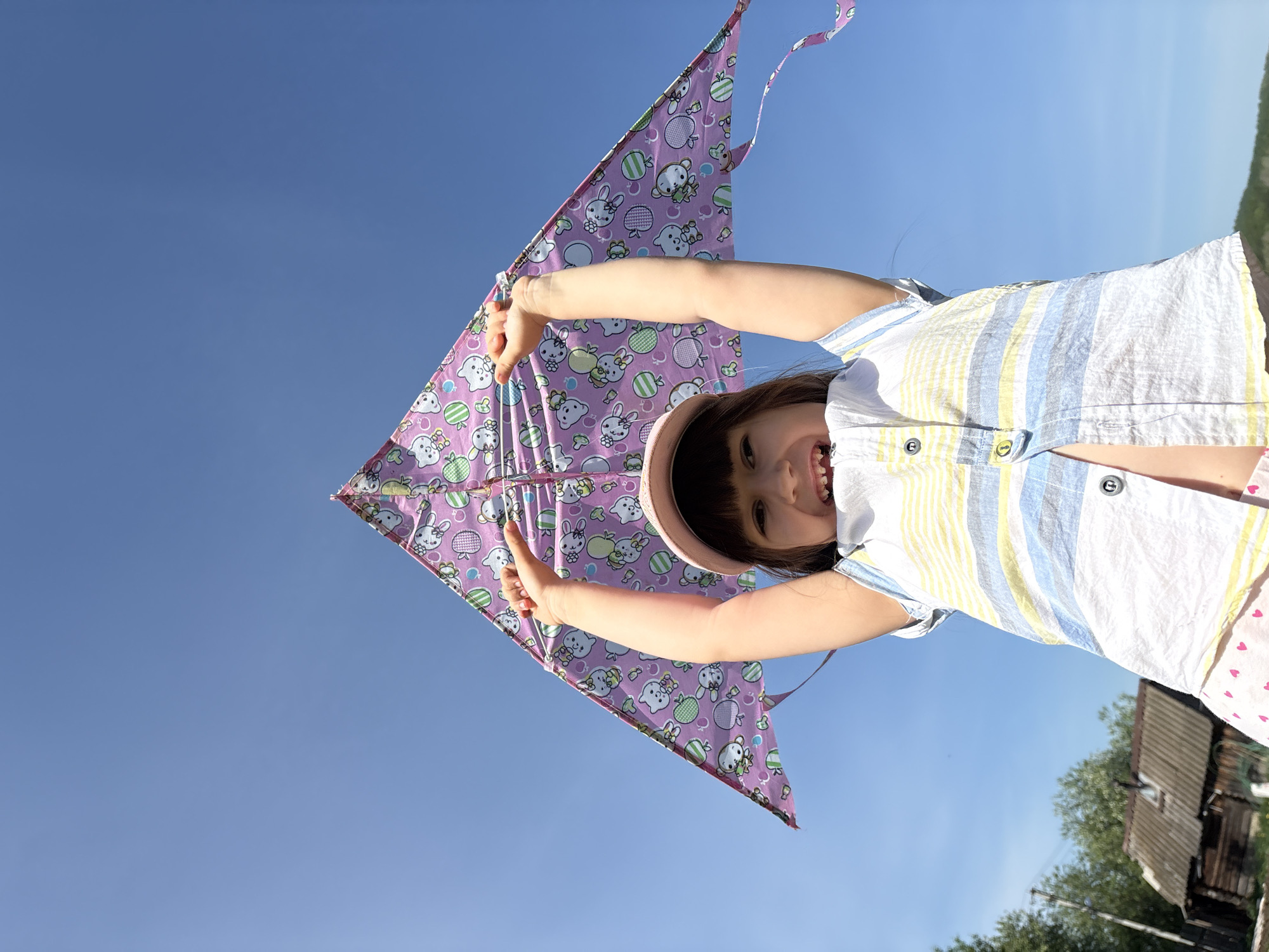A young girl is flying a kite on a clear day, with a blue sky background. The girl is holding the kite above her head with both hands, smiling broadly. The kite is pink and patterned with cartoon-like animals. The girl is wearing a light-colored striped shirt and a pink hat. The scene is outdoors, with some trees and a building visible in the background, in Nizhnyaya Chermoda, Russia.