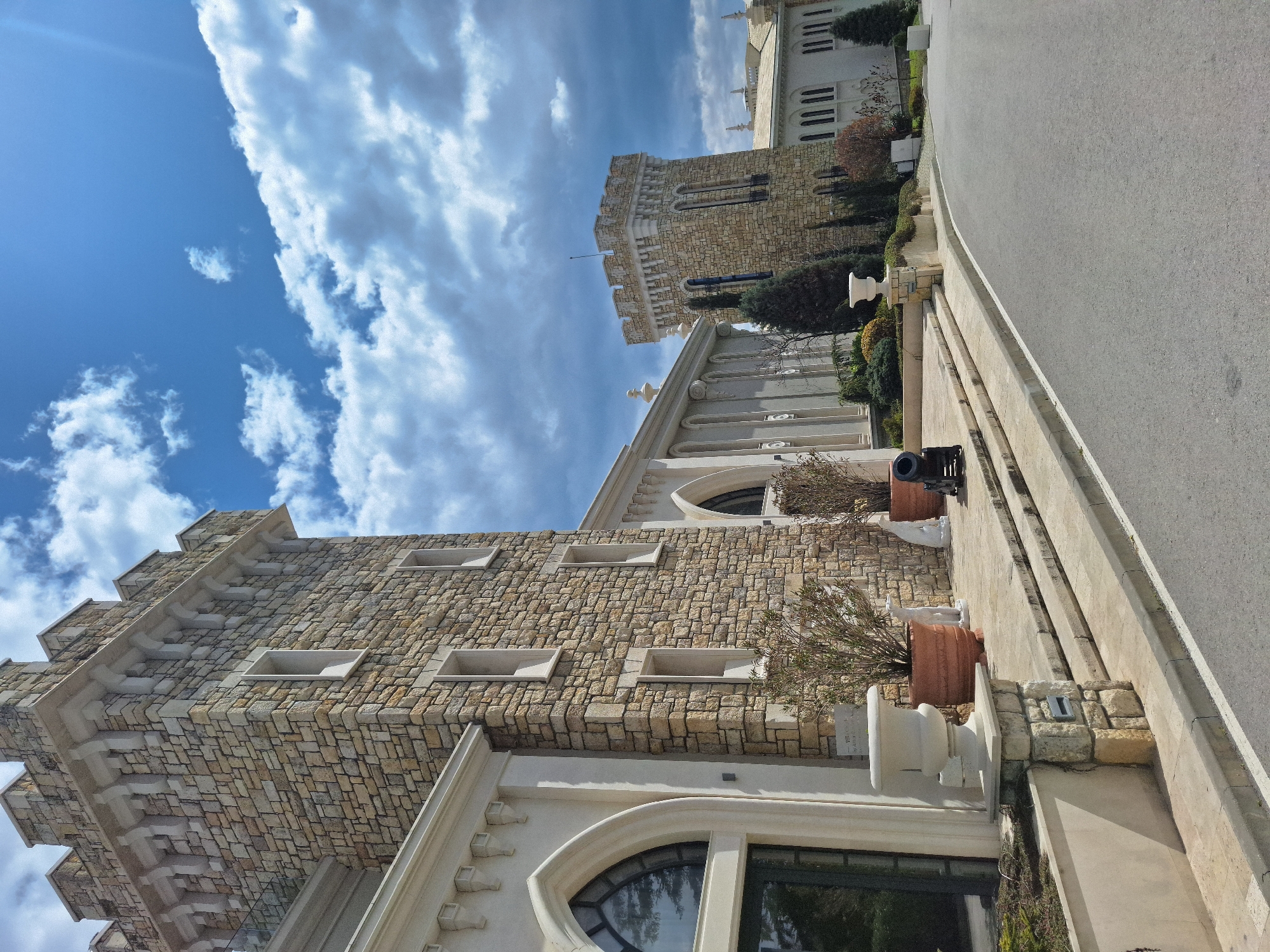The image depicts an architectural scene in Varna, Bulgaria. The scene features buildings constructed of stone, one of which includes a tower. The sky is blue with white clouds. The composition includes a paved roadway and ornamental features such as potted plants and a cannon. The buildings exhibit a mix of textures and decorative elements, reflecting a specific architectural style. The image captures a portion of the built environment within the city, highlighting its unique aesthetics.