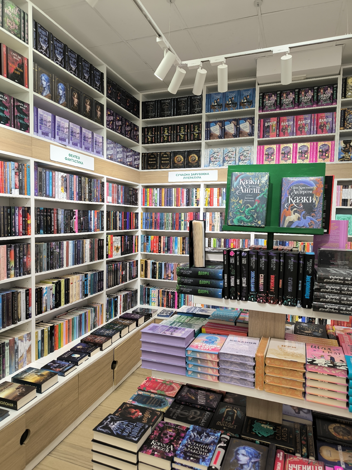 The image depicts the interior of a bookstore in Vinnytsia, Ukraine. Shelves are stocked with a wide variety of books, categorized by genre and author. Signage indicates sections for "Fantasy" and "Contemporary Foreign Literature." A central display table features stacks of books with different colored covers, including purple, orange, and teal. Many of the books are in Ukrainian. The lighting is bright and modern. The floors are light-colored wood. The scene is organized and well-stocked.