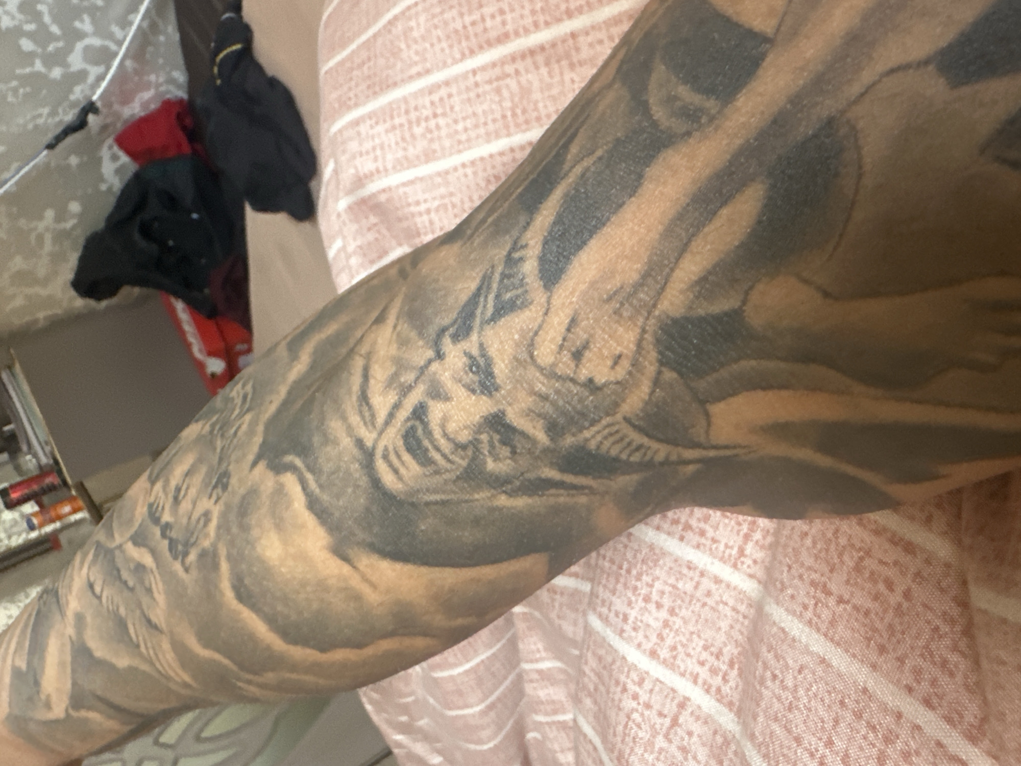 A close-up shot of a tattooed arm with a design featuring an animal and wing-like elements, resting against a horizontally striped fabric. To the left of the arm, in the background, is a jumble of dark clothing and items that might be on a rack or shelf. The environment is indoors. The location is unknown.
