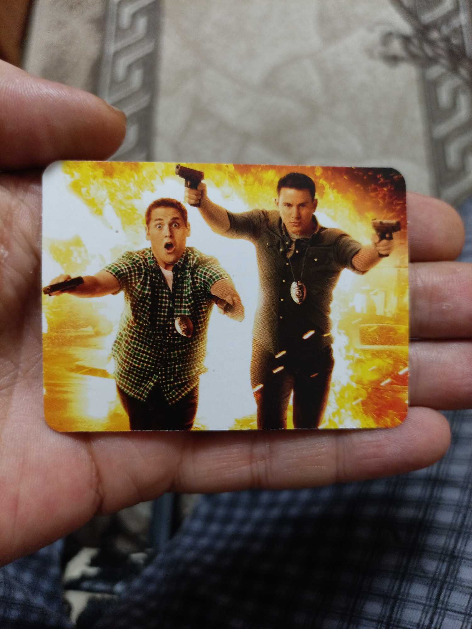 The image is a close-up of a rectangular card held in a person's hands. The card features an action movie scene with two men holding firearms. The man on the left is wearing a green and black plaid shirt and has a surprised expression, holding a gun in his right hand. The man on the right is wearing a dark shirt and is also holding a gun in his right hand, with a badge hanging from his neck. Both men appear to be running through a fiery background. The card has rounded corners. The background of the image is out of focus and appears to be a textured surface, possibly fabric or wallpaper, in muted tones. There is no indication of the city of Nyagan, Russia, or any identifiable landmarks within the image.