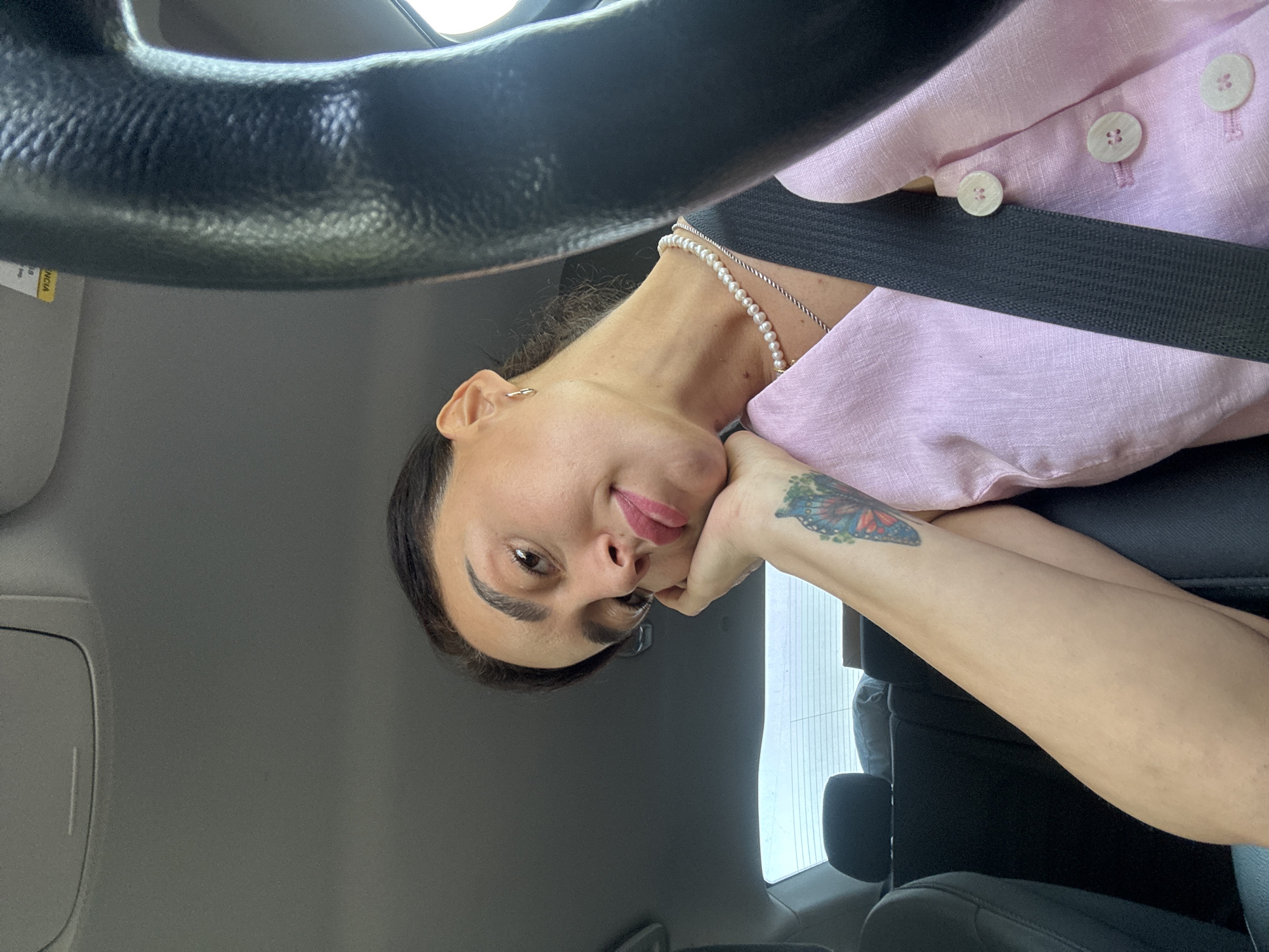 A woman is pictured inside a car, likely in Tbilisi, Georgia, given the implied context. She is resting her head on her hand, with her left arm featuring a butterfly tattoo. She is wearing a light pink top with pearl buttons and a pearl necklace. A black seatbelt is visible across her chest. The steering wheel of the car is in the foreground on the right side of the image. The background shows the interior of the car, including the back seats and the rear window.