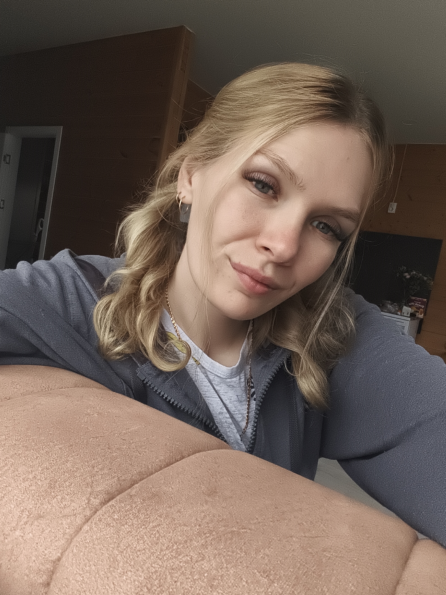 A young woman with blonde hair and blue eyes is taking a selfie indoors, leaning on a textured orange couch. She is wearing a grey zip-up hoodie over a grey t-shirt and a delicate chain necklace. The background includes a dark wood paneled wall and a doorway. The lighting suggests it is daytime. The location is described as "Русский Вожой, Russia" which translates to "Russian Vozhoj, Russia."