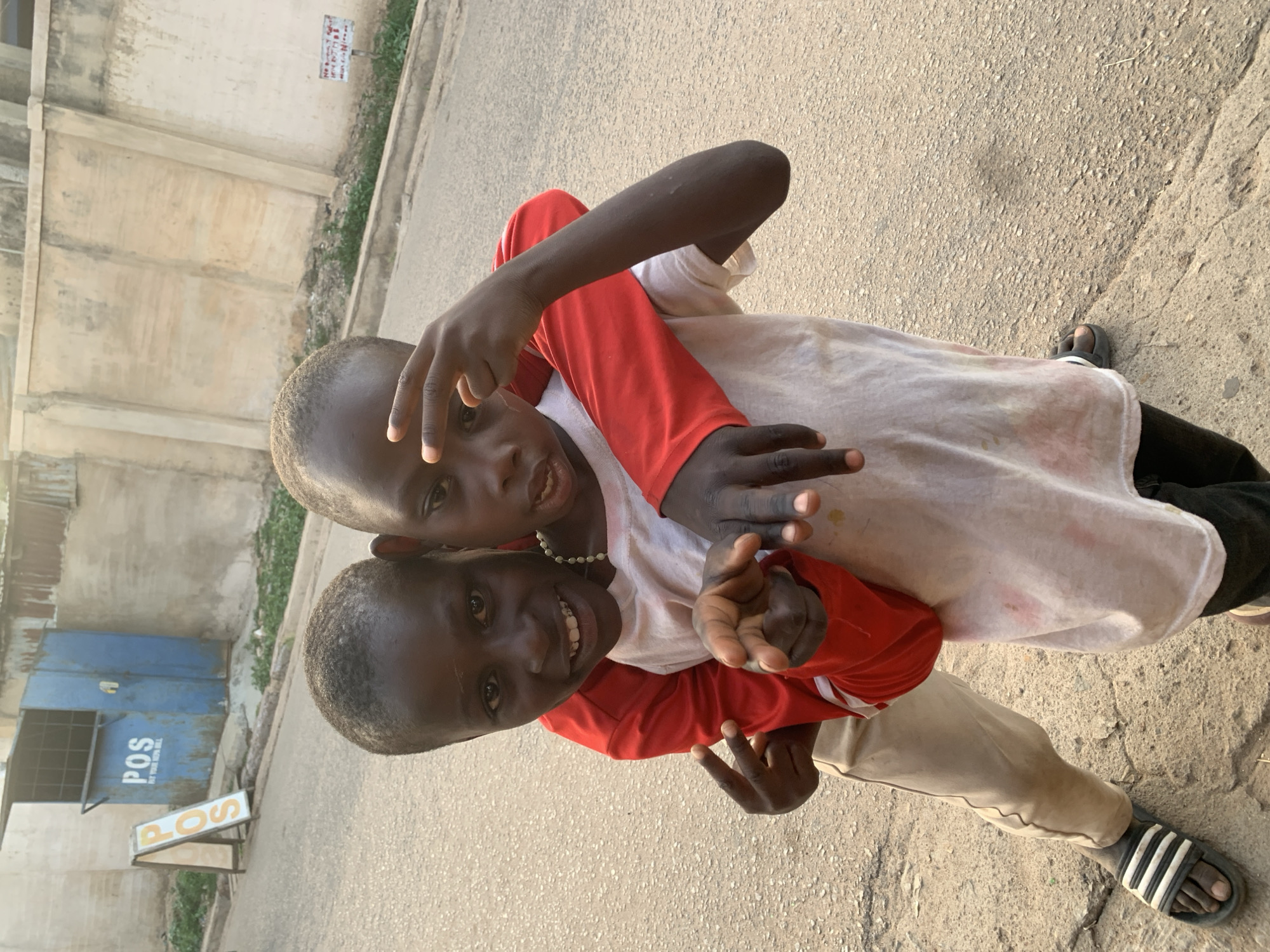 Two young Black boys, one giving the other a piggyback ride, pose for a photo outdoors on a cracked pavement. The boy on top wears a white t-shirt over a red long-sleeved shirt, black pants, and dark sandals, and is making a peace sign with his left hand. The boy carrying him wears a red long-sleeved shirt over a white t-shirt, light brown pants, and Adidas sandals, and also flashes a peace sign. They are smiling at the camera. In the background, there is a light-colored building, a blue metal POS kiosk, and a sign reading "POS" with "At Your Service 24/7" below. Based on the presence of the POS kiosk, the location is possibly Jos, Nigeria. The time of day appears to be mid-day.