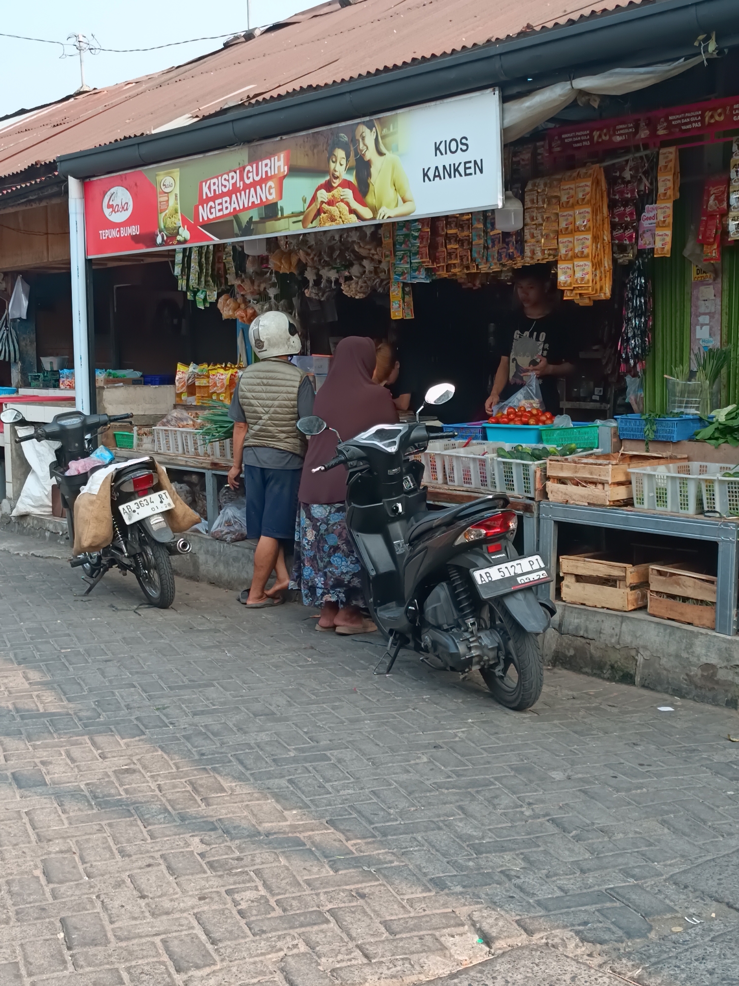 The image captures a street-level view of a small shop in the city of Yogyakarta, Indonesia. The shop appears to be an outdoor market stall or a small convenience store. It is adorned with a red and white sign that reads "KRISPI, GURIH, NGEBAWANG" and features an image of a person eating fried chicken. Another sign, white with black lettering, says "KIOS KANKEN". The shop sells various food items, including packaged snacks and fresh produce such as tomatoes and cucumbers, displayed in baskets and crates.
In the foreground, two motorcycles are parked on a cobblestone street. One motorcycle, a black scooter, is in the center of the image, with two burlap sacks attached to its rear. A license plate on this scooter reads "AB 3634 RT". The second motorcycle, also a black scooter, is to the right, partially visible.
Three individuals are present in the scene. One person, wearing a white helmet and a khaki vest over a grey shirt and blue shorts, stands near the motorcycle. Another person, wearing a brown hijab and a patterned skirt, stands to the right of the first individual. A third person, a young male wearing a black t-shirt with a graphic print, is behind the counter of the shop, holding a plastic bag. The environment is typical of a local marketplace, with goods displayed openly. The sun appears to be shining, casting shadows on the cobblestone ground. The overall impression is that of a bustling local retail scene.