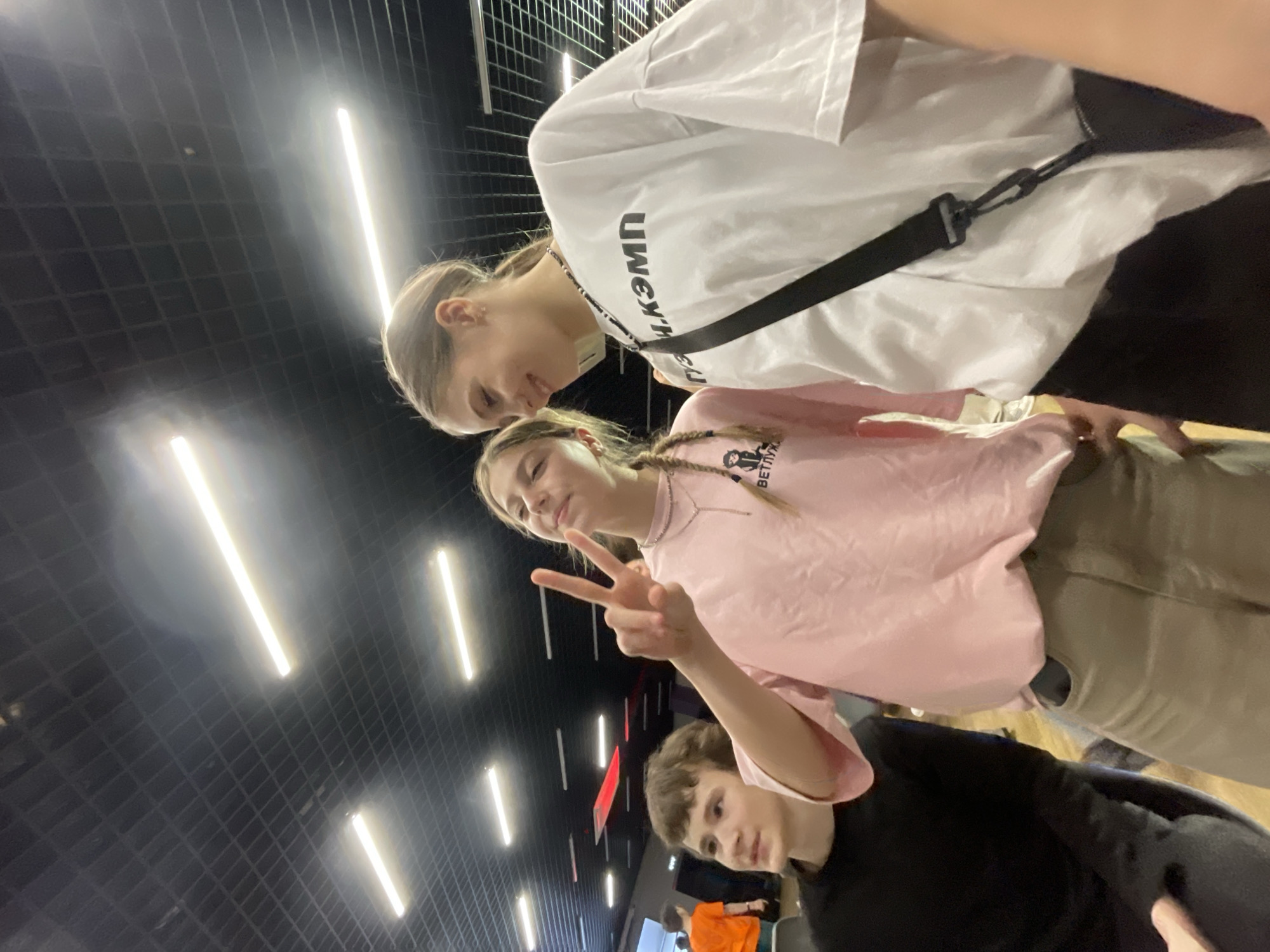 An indoor scene in a modern, brightly lit facility features three young individuals in the foreground and midground. In the left-center foreground, a young female with light brown braided hair wears a pink short-sleeved t-shirt with a graphic that includes a cartoon character and the Cyrillic text "ВЕТЛУЖА" (VETLUZHA). She also wears a silver-colored chain necklace with a pendant and khaki-colored pants. Her right hand is raised, forming a "peace" sign, and she displays a slight smile, looking directly towards the camera. To her right, in the right foreground, a second young female with light brown hair pulled back wears a white short-sleeved t-shirt with the Cyrillic text "ГУМ.КЭМП" (GUM.KEMP) in black across the chest. She has a black beaded necklace and a black strap from a cross-body bag across her torso. Her head is tilted, and she is smiling. In the left background, a young male with short brown hair, wearing a black long-sleeved shirt, looks off to his left. The background features a dark, grid-patterned ceiling with multiple bright, linear LED light fixtures. A red rectangular illuminated panel is visible on the upper left wall, and a large, light-colored rectangular screen or panel is partially visible in the far left background. Other individuals are indistinctly visible further in the background. The scene is consistent with the interior of a youth activity or educational center.
Based on the visible text "ГУМ.КЭМП" (GUM.KEMP) on one individual's t-shirt and the modern interior suited for youth programs, the location is identified as the **Центр развития личности "ГУМ.КЭМП" (Center for Personality Development "GUM.KEMP") at Ulitsa Maerchaka, 60, in Krasnoyarsk, Russia.**