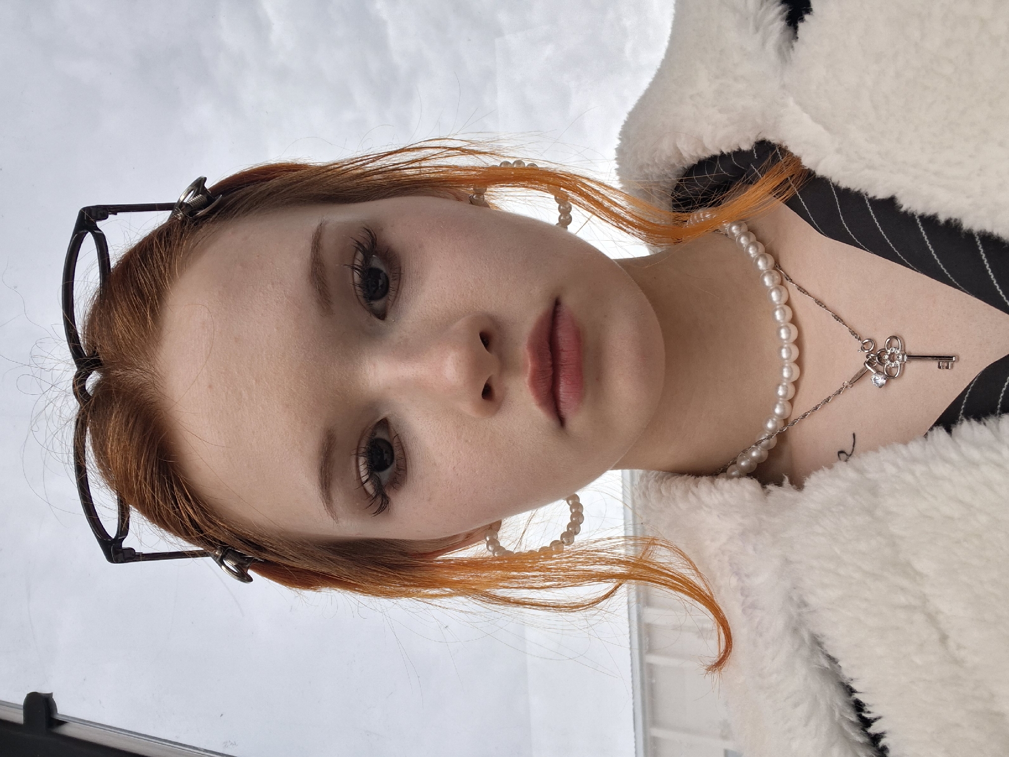 A young woman with pale skin and auburn hair is posing for a selfie. She is wearing eyeglasses perched on her head, pearl earrings, a pearl necklace with a key pendant, and a fluffy white jacket over a black and white striped top. A small tattoo is visible on her collarbone. The background appears to be a cloudy sky visible through a window. The location is possibly Chaikovsky, Russia.