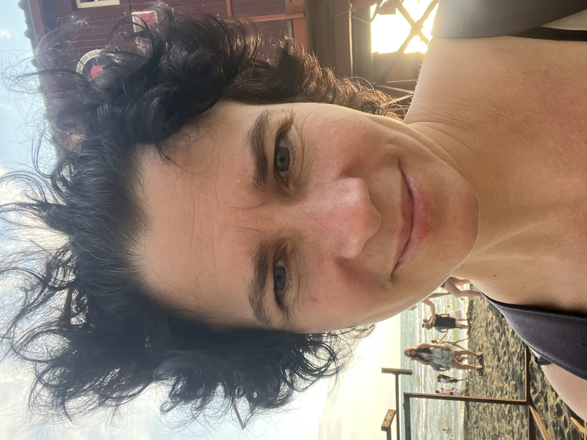 A selfie of a woman with dark, curly hair and fair skin is taken on a beach in Ureki, Georgia. The woman is in the foreground, facing the camera and smiling slightly. Her hair is windblown, and she appears to be wearing a dark top. Behind her, the beach is sandy with people walking and standing near the water. The Black Sea is visible in the background, with gentle waves. The sun is setting, casting a warm glow on the scene and creating lens flare. A wooden structure, possibly part of a lifeguard stand or beach facility, is visible on the right side of the image. The overall atmosphere is relaxed and summery.