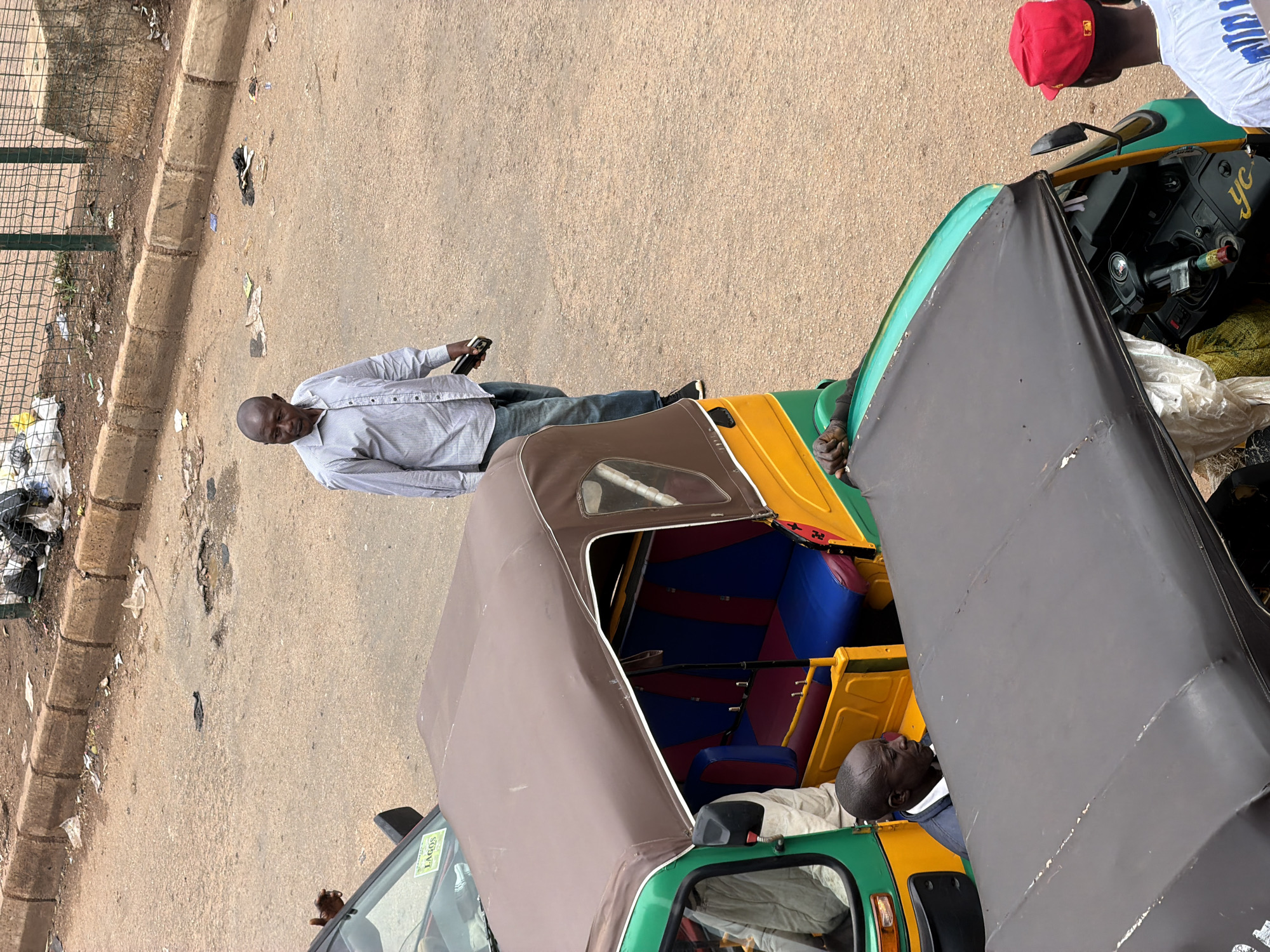 The image captures a street scene in Bukuru, Nigeria. In the foreground, two auto-rickshaws are partially visible. The auto-rickshaw on the left is predominantly green and yellow, with a brown canopy covering its passenger area, which features red and blue seating. A man in a dark jacket and white shirt is seated inside the driver's compartment. To the right of this auto-rickshaw, another auto-rickshaw is mostly obscured by the first, with only its black canopy and some of its driver's area visible. A man in a red baseball cap and white t-shirt is standing to the right of this second auto-rickshaw. In the midground, a man in a light blue, pinstriped shirt and blue jeans is walking away from the camera, holding a black mobile phone in his right hand. The background shows a dusty, paved road with a concrete curb along the edge, and beyond that, a metal fence and some debris. The overall impression is of a typical street scene in a Nigerian city.