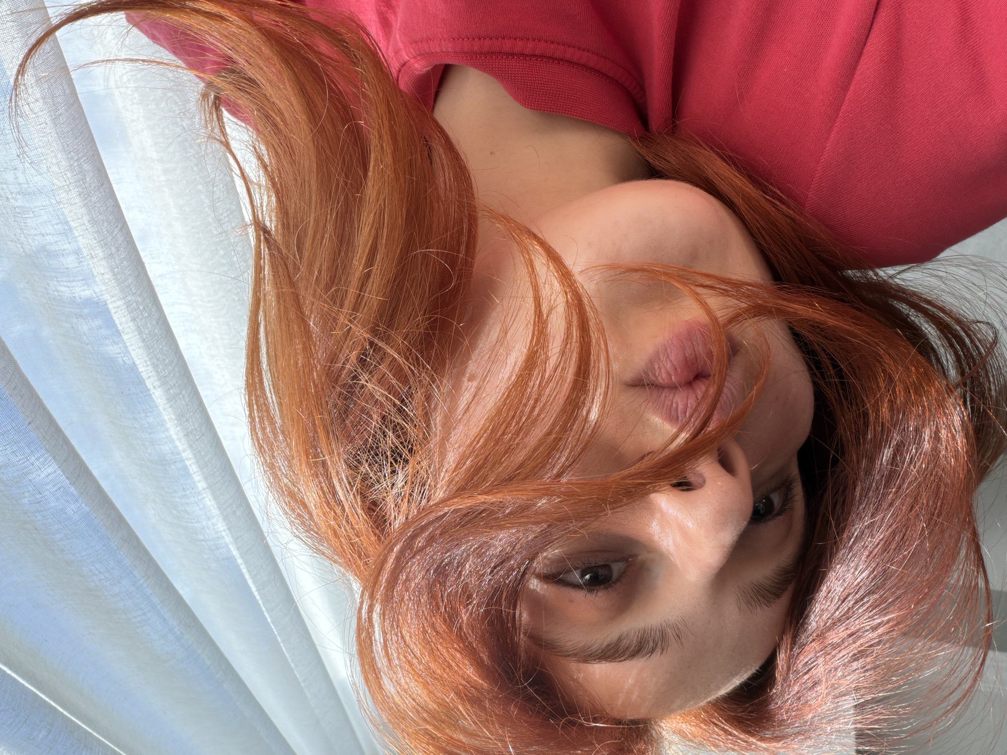 The image shows a close-up, upside-down perspective of a person's face, likely a woman, with vibrant red hair. She is wearing a red top. Her face is well-lit. A curtain or pleated blind is visible in the background. It is not possible to determine the location as Tbilisi, Georgia without additional information.
