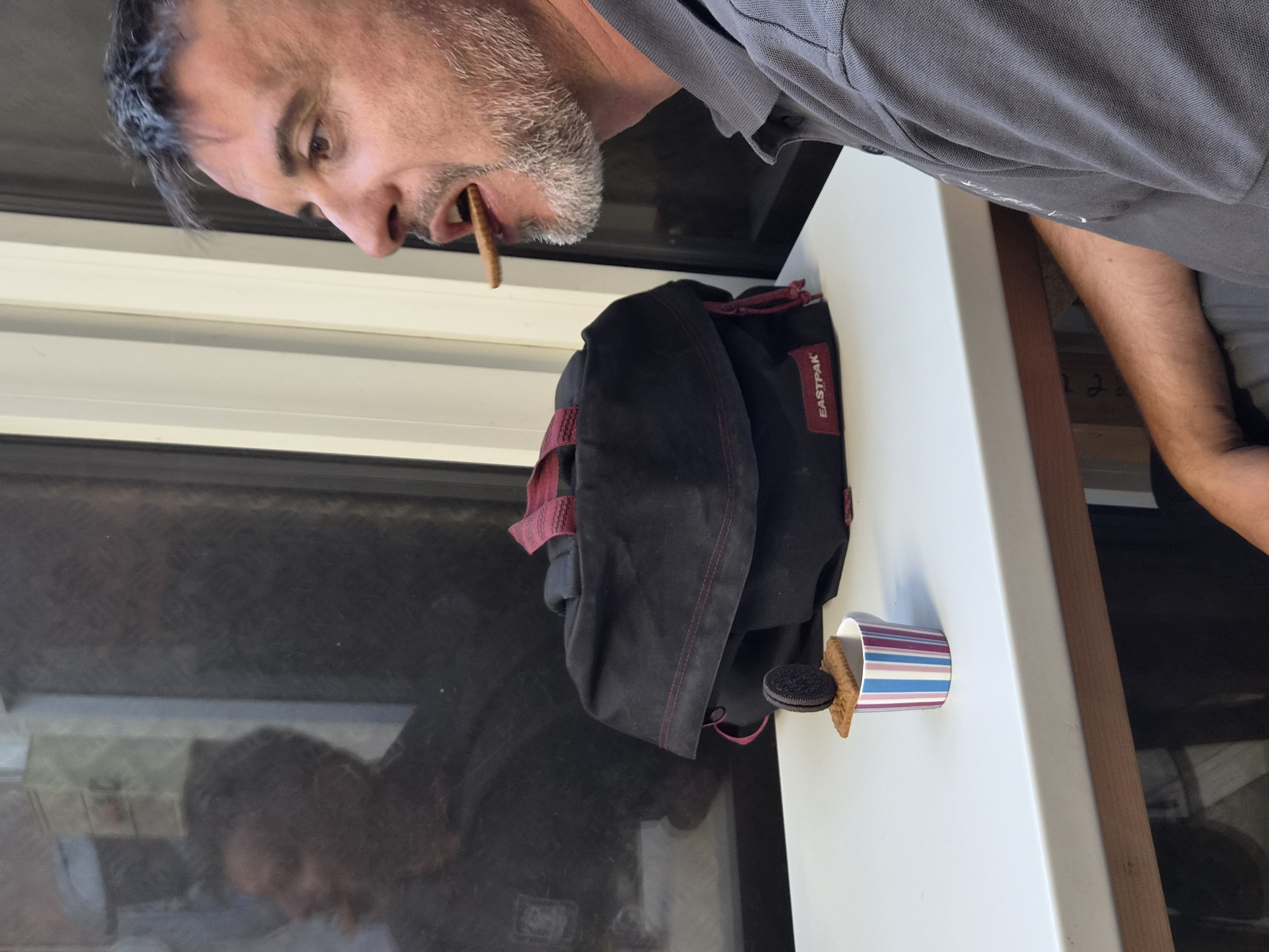 A man is sitting at a white table, with a black Eastpak backpack behind him. He is eating a biscuit that is in his mouth and on top of a biscuit and an Oreo cookie which are stacked on a striped cup. The man has gray hair and a beard and is wearing a gray polo shirt. He is looking to his right, towards a window. There is a reflection in the window of another person and part of the interior of a vehicle, possibly a train or bus. The scene appears to be taking place within a moving vehicle or a place with a large window, consistent with a travel context in Leudelange, Luxembourg.