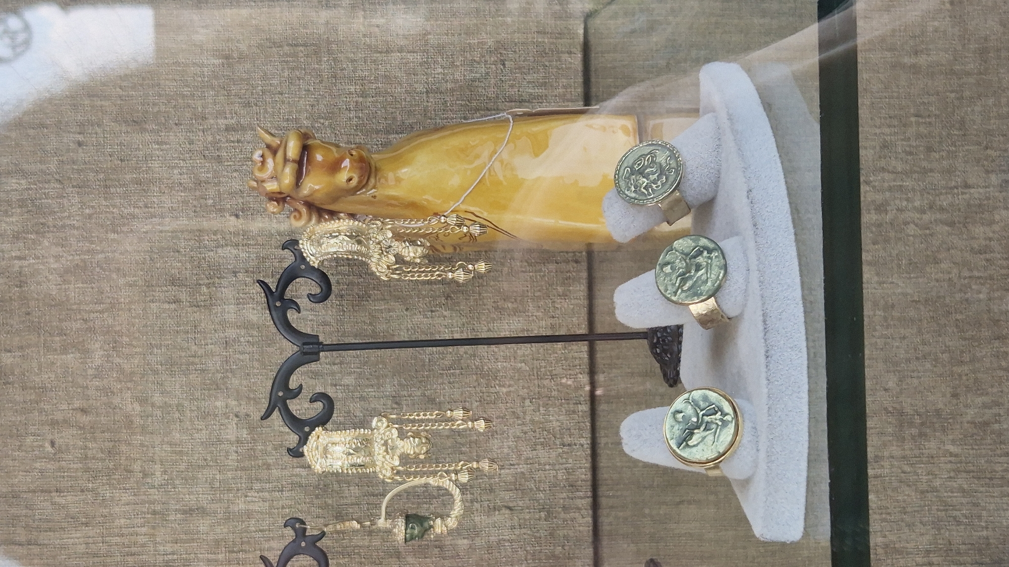 A window display in Taranto, Italy, showcases a collection of jewelry. In the upper portion of the display, two ornate gold chandelier-style earrings hang from a black metal stand. To the right of the earrings, a shiny yellow ceramic horse figurine stands prominently. Below, three rings are arranged on a light grey velvet display. Each ring features a circular, greenish medallion with an embossed design, likely depicting historical figures or mythological scenes, set in a gold band. The background of the display is a textured burlap-like material. The overall scene is well-lit, highlighting the detail and craftsmanship of the jewelry and decorative items.