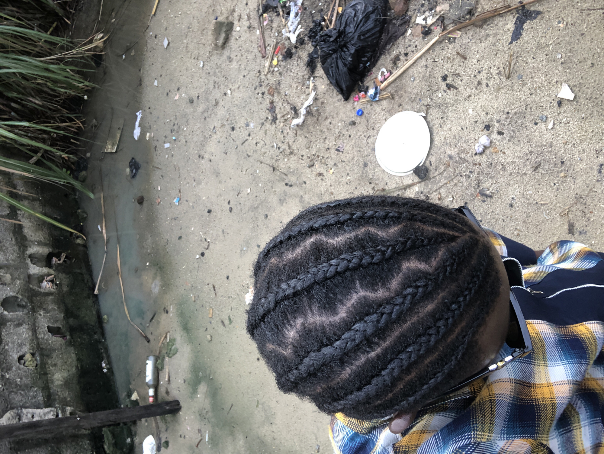 A person with braided hair is viewed from above, wearing a plaid shirt. They are standing on a sandy ground littered with trash, including plastic bags, bottles, and debris. In the background, there is a concrete wall and some water with more trash. The scene appears to be in a neglected area.