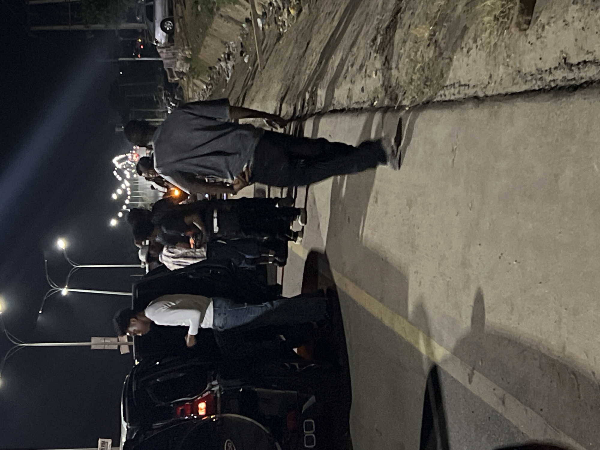 The image shows a group of people gathered on a street in Abuja, Nigeria, at night. Several individuals are standing near a dark-colored vehicle with its trunk open. Some of them are looking into the trunk. Streetlights illuminate the scene. One man is walking away from the group. The background shows an area with waste and other debris.