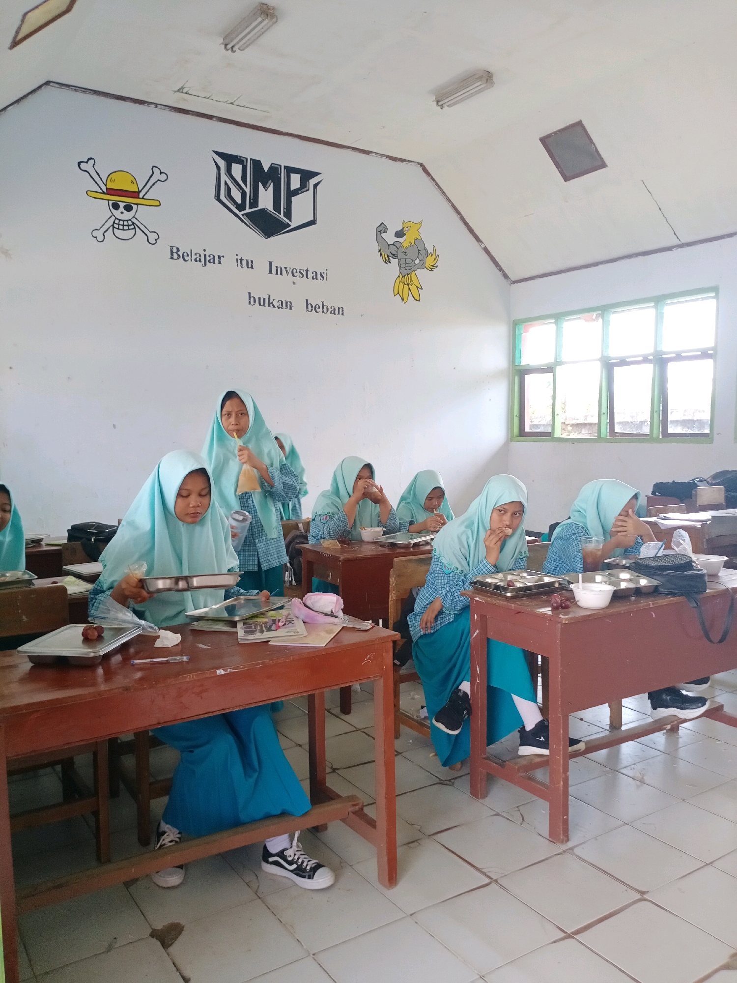 The image depicts the interior of a classroom in Tragah, Indonesia, likely a junior high school, given the "SMP" logo on the wall. Several young female students, all wearing light blue hijabs and uniforms, are seated at wooden desks. The classroom walls are white, with a mural featuring a skull with a straw hat (reminiscent of the anime One Piece), the "SMP" logo, and motivational text in Indonesian: "Belajar itu Investasi bukan beban" (Learning is an Investment, not a burden). Another mural depicts a muscular cartoon bird. The students appear to be eating or drinking, with trays of food and drinks on their desks. One student in the foreground is holding a tray with food and a cup, while another in the background is drinking from a plastic bag with a straw. The lighting suggests it is daytime, with light coming in from a window on the right side of the room. The floor is tiled and appears worn. The desks are made of dark wood, and some have bags and other personal items on them.