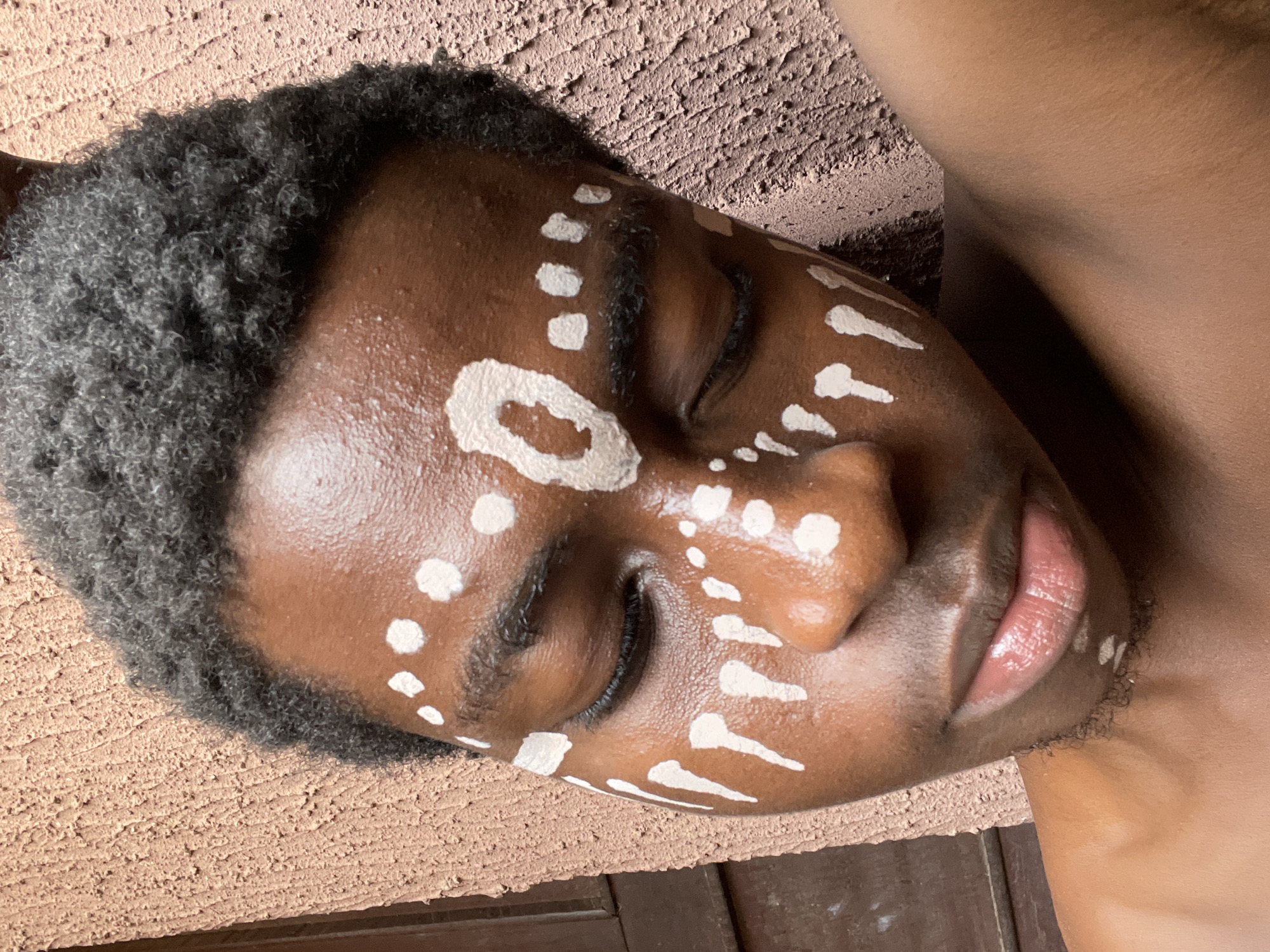 The image shows a person of African descent with closed eyes, adorned with white facial markings, resting on what appears to be a textured surface in Nkpor, Nigeria. The facial markings consist of dots, lines, and an oval shape. The background includes a rough, potentially painted surface and a dark wooden structure. The person's hair is short and dark. Their lips have a glossy appearance. Their shoulder is visible at the right edge of the frame.