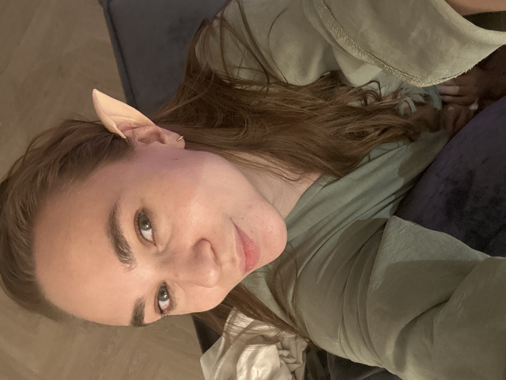 A woman is looking at the camera, wearing an elf ear. She is wearing a green shirt. She is inside, sitting on a gray couch with a blue pillow. The location of Kharnavy, Russia cannot be determined from the image.