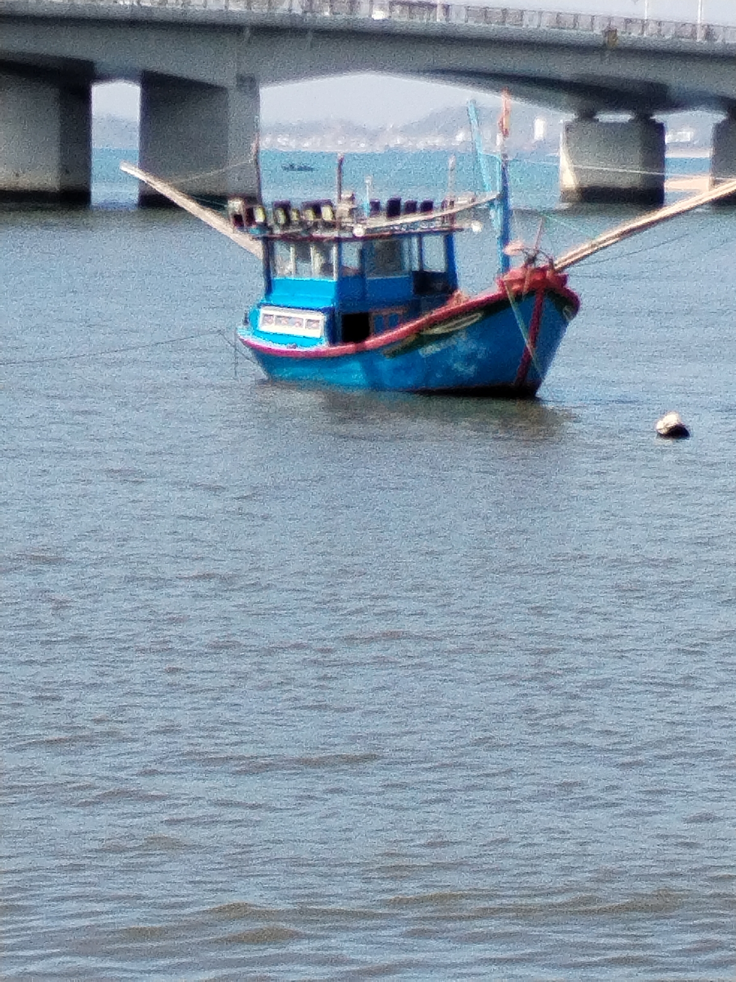 A bright blue fishing boat with red trim is anchored in calm, blue water. The boat has a cabin structure on its deck, and several lights are visible. Two wooden booms extend from the sides of the boat, angled upwards. In the background, a large concrete bridge with multiple support pillars spans the water. The bridge appears to be part of a coastal highway. The overall scene suggests a sunny day in a coastal area of Vietnam, likely in or near Nha Trang city, given the prominent bridge infrastructure and typical fishing vessel design.