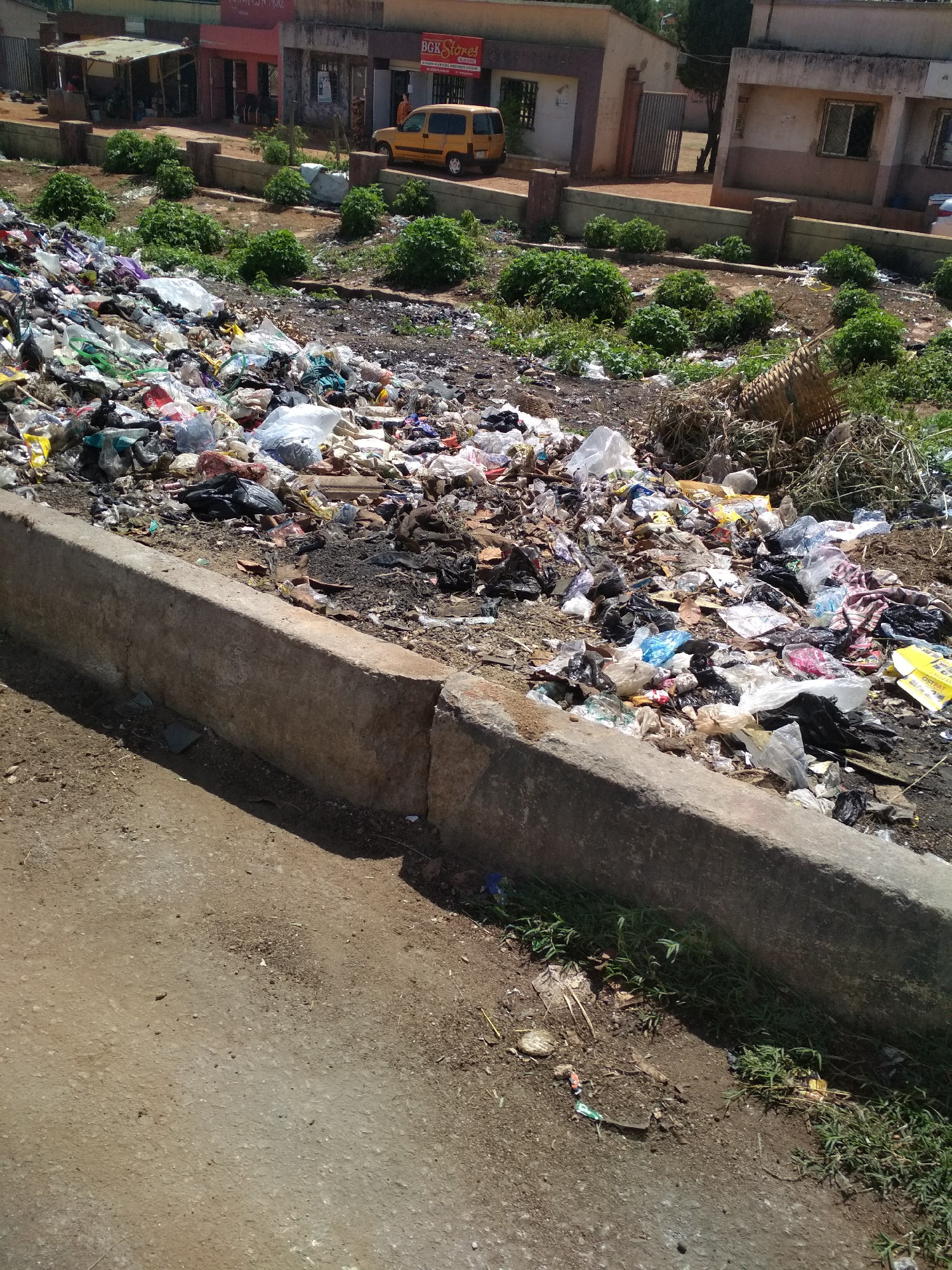 The image depicts a large pile of uncollected garbage along a street in Gura Top, Nigeria. The refuse consists of plastic bags, food waste, and other debris. A concrete curb separates the trash from the road. In the background, there are buildings and vegetation, indicating a residential or commercial area. A yellow car is parked near one of the buildings. The presence of garbage suggests issues with waste management.