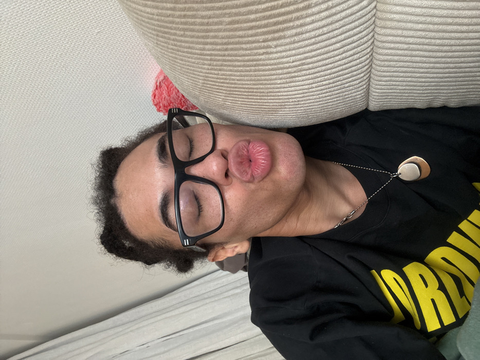 The image presents a close-up, angled portrait of an individual, likely an adult, captured from the chest up. The subject has medium-dark skin, dark curly hair styled in two small knots on top of the head, and wears black-framed eyeglasses with clear lenses. The eyes are closed, and the lips are puckered in a "kissing" pose. The subject wears a black long-sleeved t-shirt with yellow lettering partially visible on the chest. Two necklaces are worn: a silver-toned chain with varied links, and a ball chain with a layered pendant featuring a larger copper-toned irregular shape beneath a smaller silver-toned irregular shape. The subject's right cheek is pressed against a beige, vertically ribbed upholstered surface, consistent with a sofa or armchair. To the left in the background, a light-colored, textured wall and a pleated curtain are visible. A small, reddish fabric item is partially obscured behind the subject's head. The scene is indoors. No specific visual cues within the image confirm the location in the city of Paris, France.