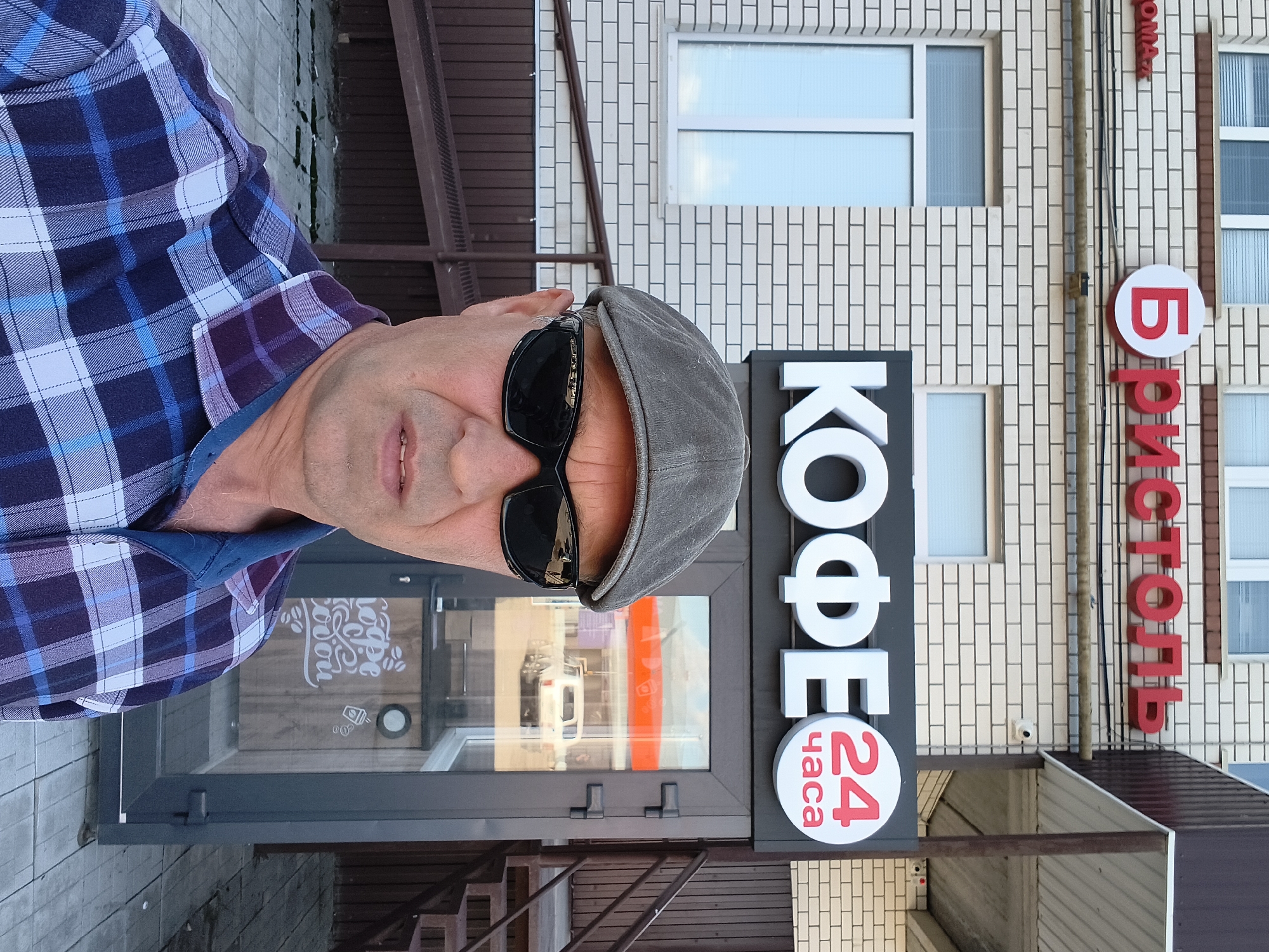 A middle-aged man wearing sunglasses and a grey flat cap, and a blue and purple plaid shirt stands in front of a coffee shop in Mikhaylovka, Russia. The coffee shop has a sign that reads "КОФЕ 24 ЧАСА" in large white letters. The building is made of light-colored bricks and has a ramp leading up to the entrance. Inside the coffee shop, a white SUV is visible through the glass. The man is taking a selfie, with his face and chest filling the foreground of the image.