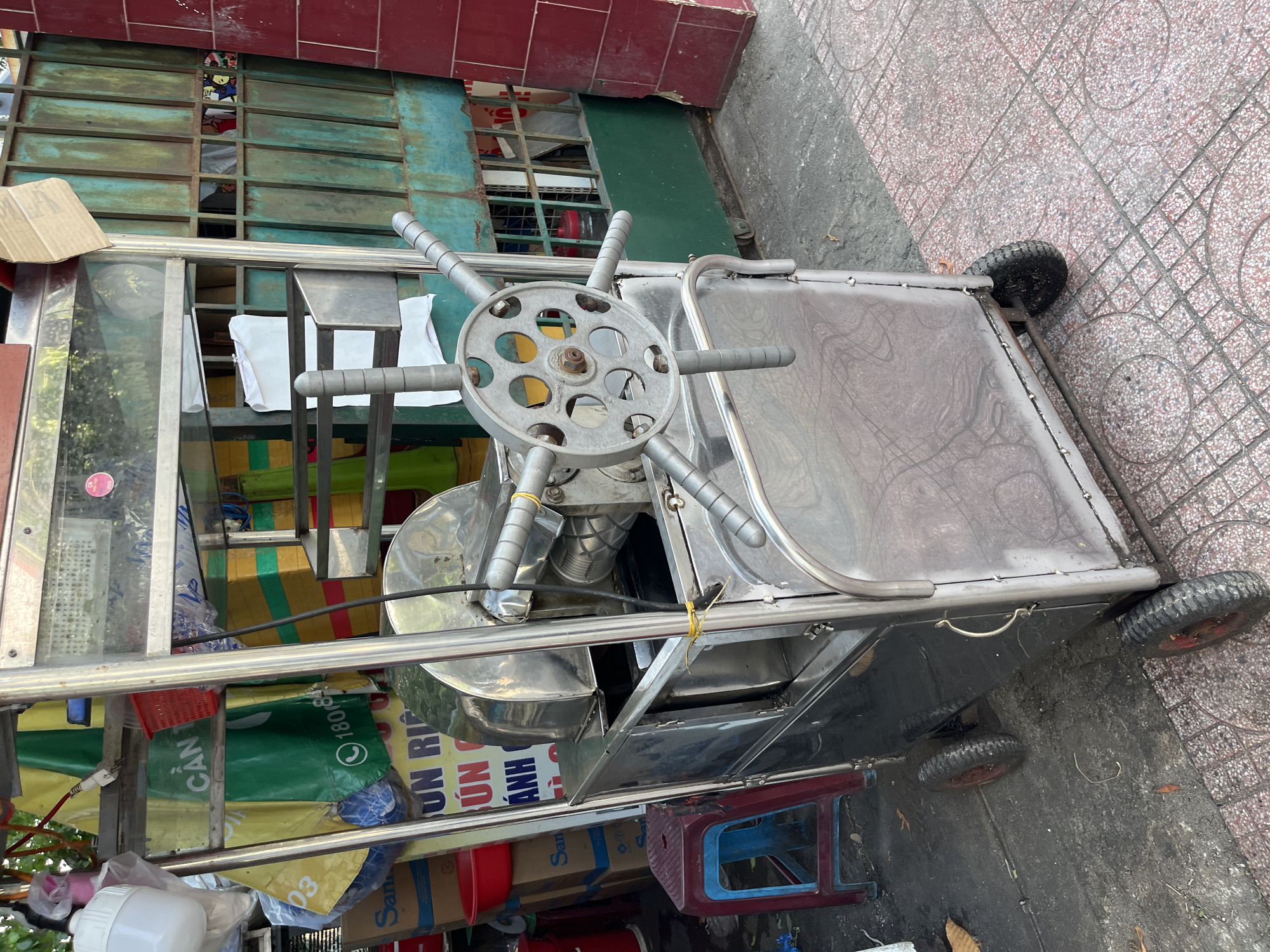 The image shows a metallic cart with a wheel in the middle, likely a food cart, located in Nha Trang Ward, Vietnam. The cart is parked on a street with patterned tiles. Behind the cart, there is a building with a green metal frame window. The cart has various items stored underneath it, including boxes and plastic bags. A small stool is located beside the cart.