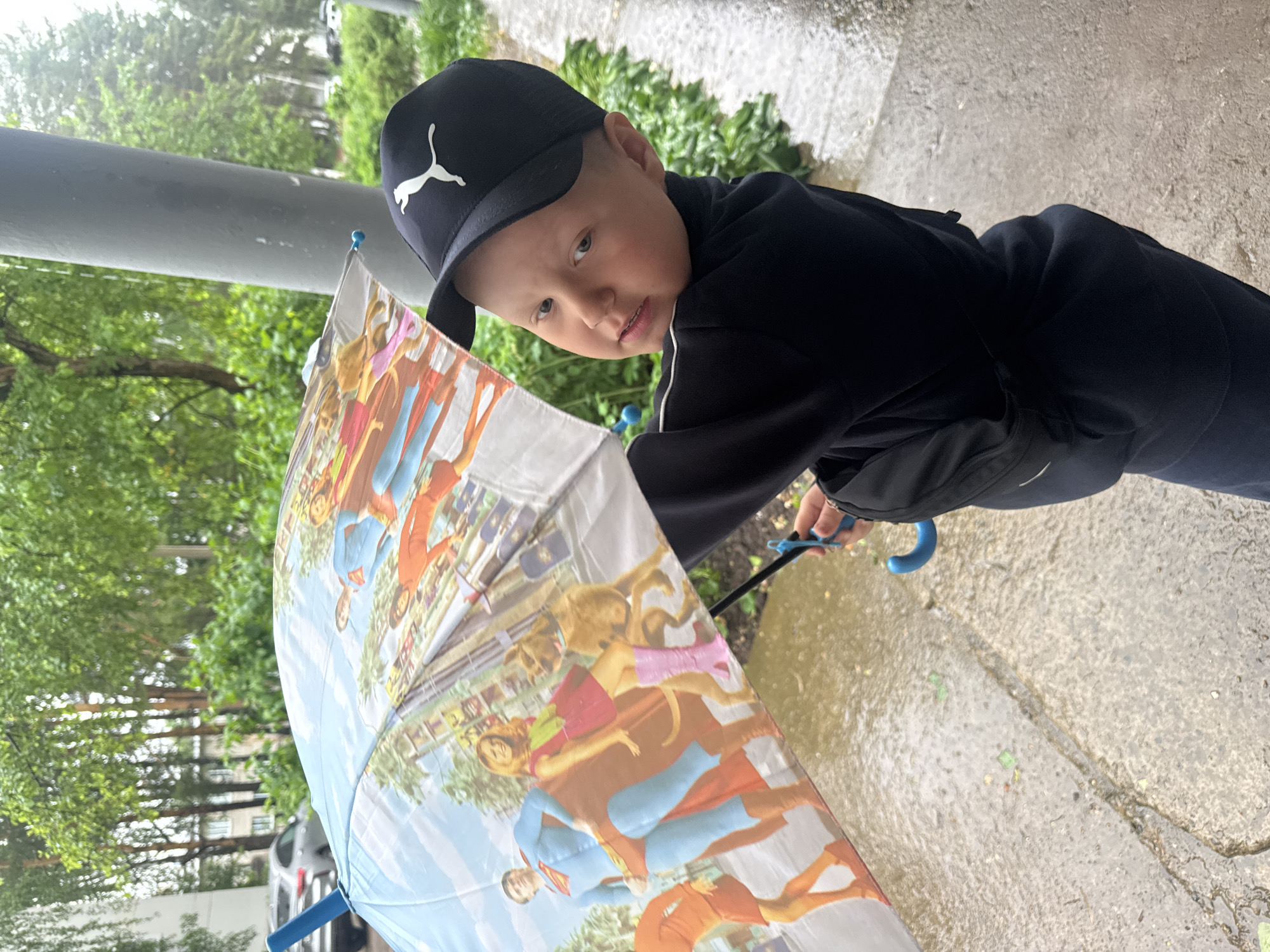 A young boy wearing a black Puma cap and a black jacket stands on a wet concrete sidewalk, holding a partially opened umbrella with images of Superman and Scooby-Doo characters. The location appears to be an outdoor area in Chaikovsky, Russia, evidenced by the urban environment. The setting includes greenery, possibly trees and bushes, visible behind the boy and to the side. A gray pole is present.