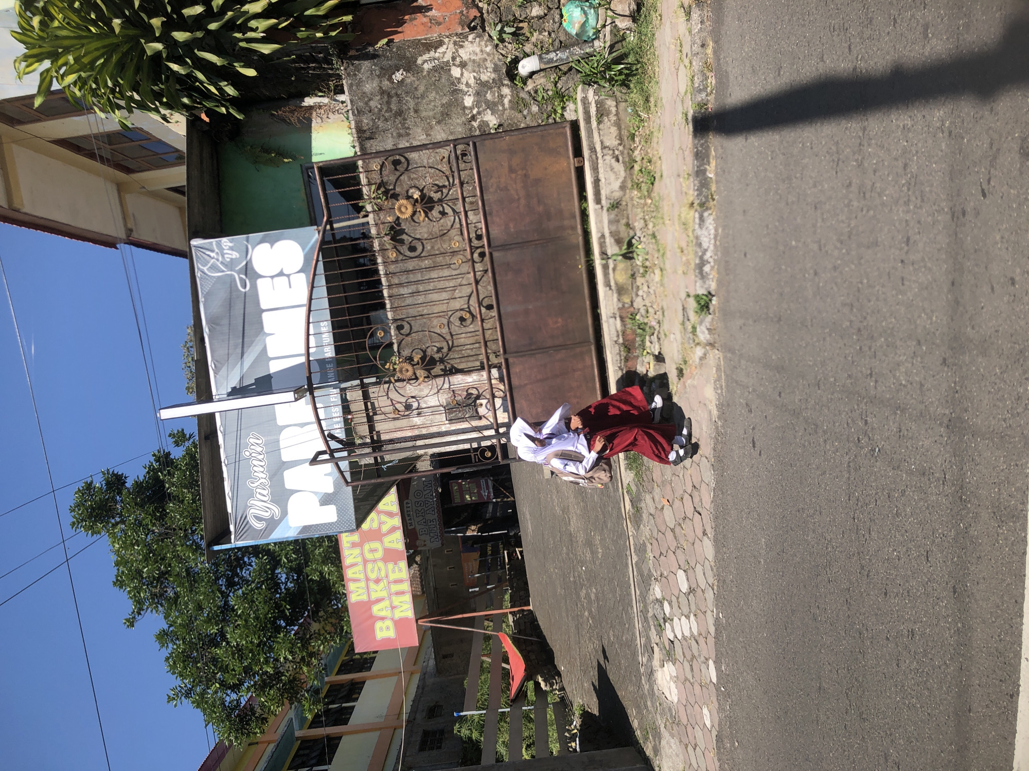 In Trawas, Indonesia, a bright, sunny day is depicted with a clear blue sky overhead. The scene focuses on a paved street with a cobblestone sidewalk. On the sidewalk, two young girls in school uniforms, complete with hijabs and red skirts, walk side-by-side. One girl carries a backpack. They are walking away from a rusty, ornate metal gate.
Behind the gate, a building with peeling paint and a green wall is visible, along with lush green trees. A large banner hangs above the gate, featuring the words "Yasmin" and "PARFUMES" in large, white lettering. To the left of the gate, a red banner announces "MANTEP BAKSO MIE AYAM" (Delicious Meatball and Chicken Noodle). The environment suggests a commercial area with buildings and street vendors. The overall impression is one of everyday life in a bustling Indonesian town.