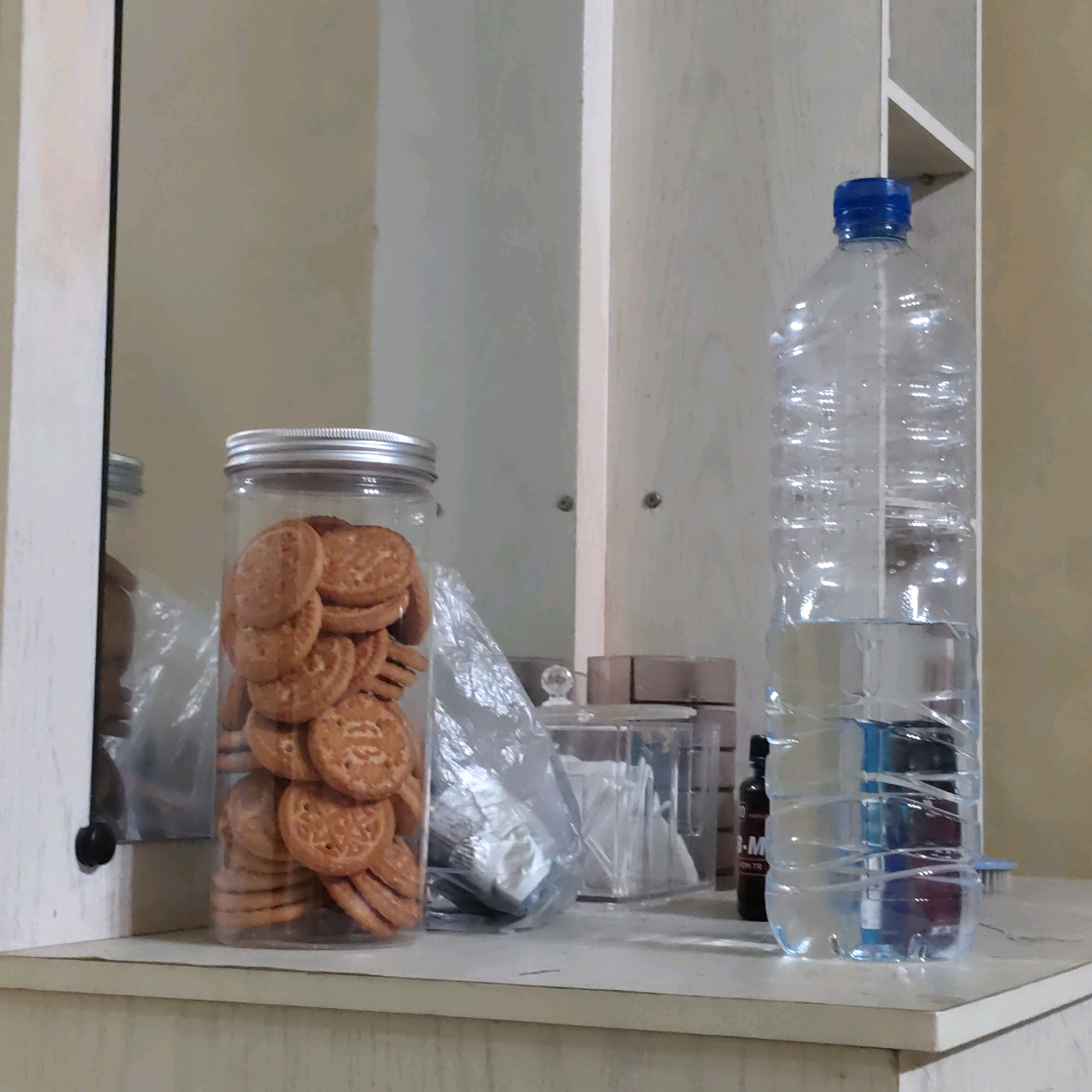 The image depicts a shelf or tabletop with several objects, set against a light-colored wall. On the left, a tall, clear plastic jar is filled with round, brown cookies. The jar has a silver lid. To the right of the cookie jar, a large, clear plastic water bottle is positioned. The bottle contains clear liquid and has a blue cap. Further to the right, a dark-colored bottle with a red label is partially visible. In the background, there appears to be a mirror reflecting some of the objects, and a white, vertical structure, possibly part of a cabinet or shelf unit. The overall scene suggests an indoor setting, possibly a kitchen or pantry. There is no visible indication of the specific location in Sunggal, Indonesia, as the image does not contain any recognizable landmarks or contextual clues for that region.