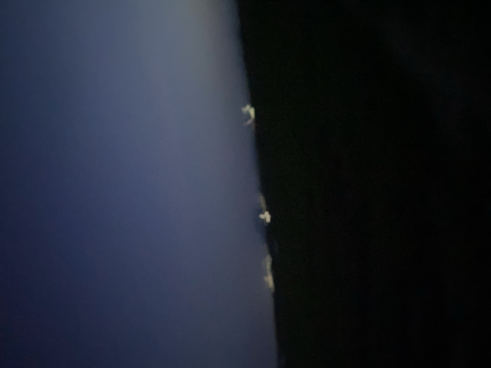 The image shows a blurry view of a dark, hilly landscape under a deep blue twilight sky. There are several indistinct, glowing white lights scattered across the horizon, suggesting distant buildings or streetlights. The foreground is almost entirely black, indicating it is a very dark, unlit area, possibly rural or undeveloped land. The general impression is one of a vast, sparsely populated area on the outskirts of a city at dusk. The specific location in Jalingo, Nigeria cannot be determined from the visual information provided in this image.