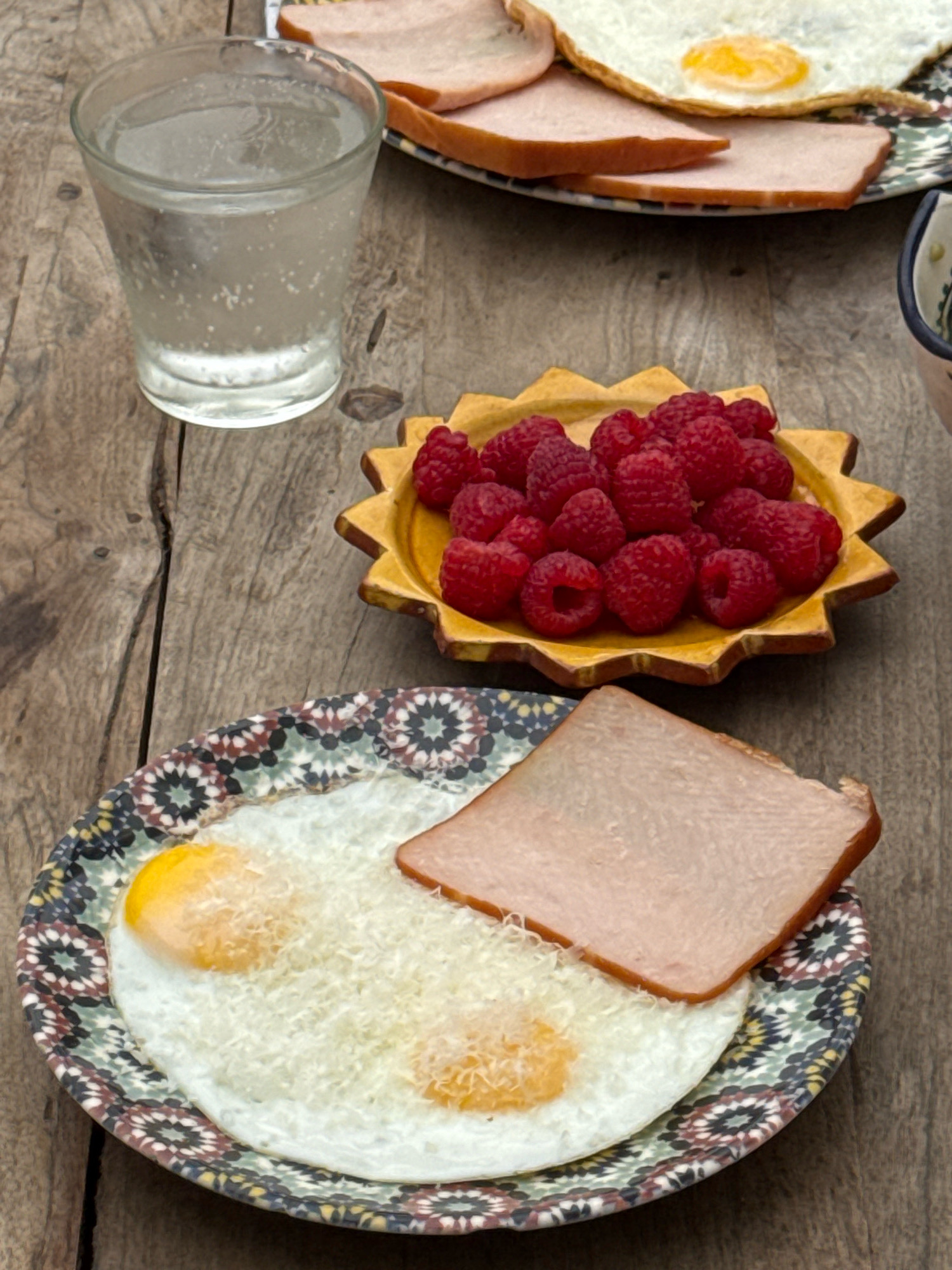 The image shows a breakfast spread on a rustic wooden table. In the foreground, a plate with two fried eggs, grated cheese, and a slice of ham is visible. To the upper left, a glass of clear liquid, likely water with ice, sits on the table. Behind the glass, another plate holds several slices of ham and one fried egg. A small, star-shaped yellow dish filled with fresh raspberries is positioned between the glass and the foreground plate. The overall setting suggests an outdoor meal, possibly on a terrace or patio. The image does not contain any specific visual cues that would allow for identification of the location as Essaouira, Morocco.