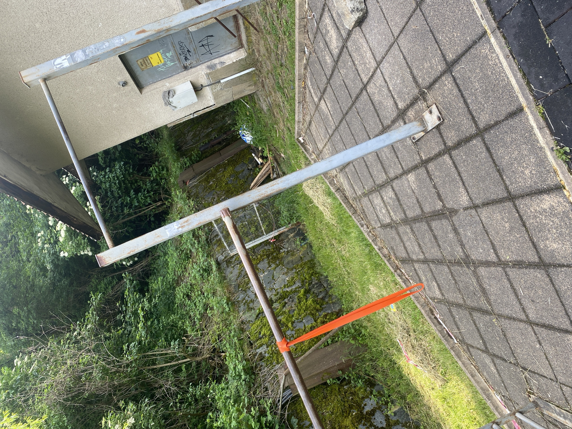 An outdoor scene shows a pull-up bar situated on a paved area next to a building and a vegetated slope. The pull-up bar is made of metal, with one horizontal bar supported by two vertical posts. An orange resistance band is attached to the horizontal bar. The building is a simple structure with a beige facade and a metal door covered in graffiti. To the left of the building and behind the pull-up bar, a steep, rocky slope is covered in moss and dense greenery. The paved area is made of square concrete tiles, with some grass and debris scattered on it. The overall impression is one of an abandoned or neglected outdoor recreational space.
