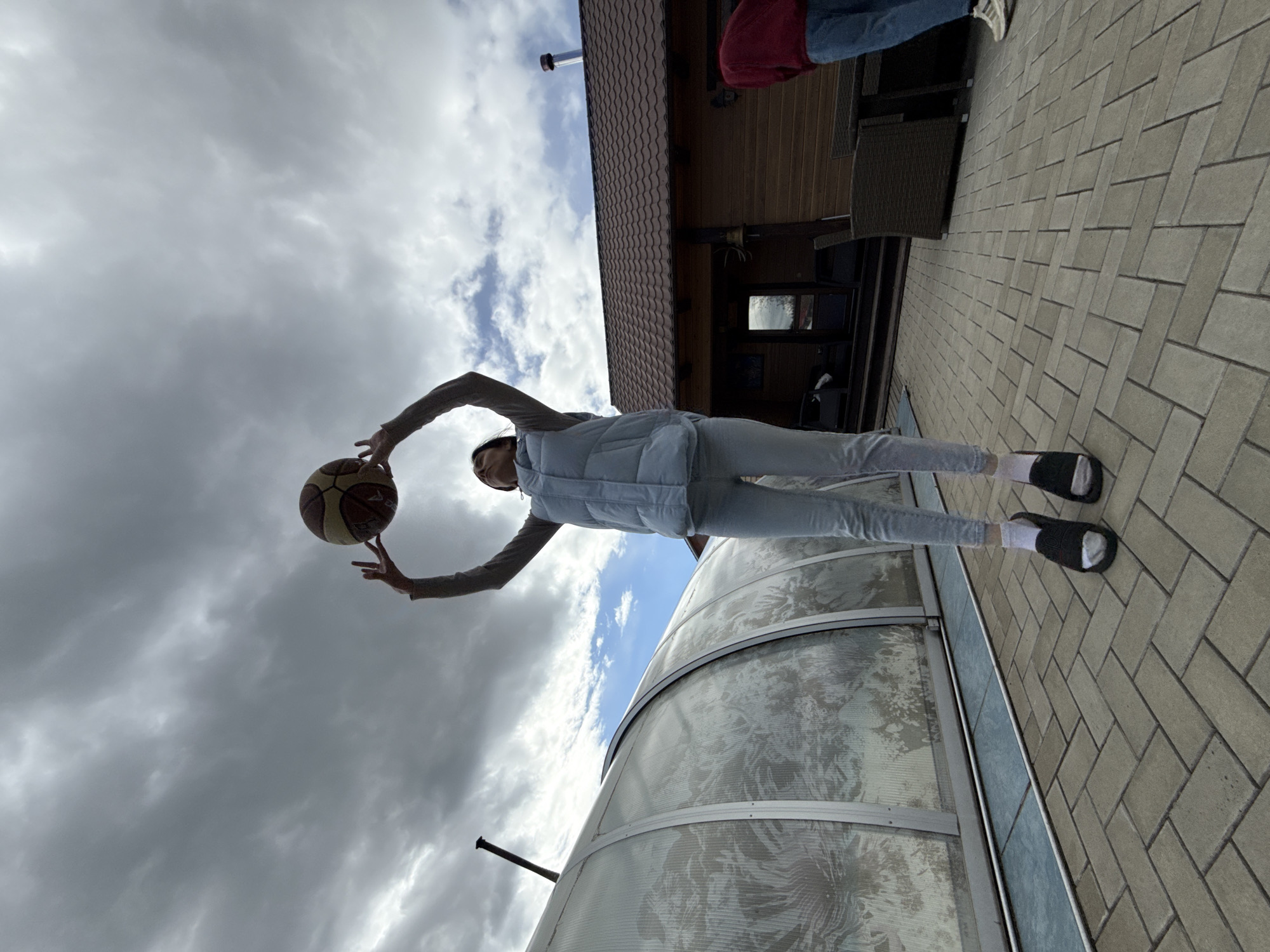 A low-angle, outdoor photograph depicts a person holding a basketball. The individual is wearing a light-colored vest over a long-sleeved shirt and light-colored pants, and dark-colored sandals with white socks. The person is positioned against a backdrop of cloudy sky. Behind and below the person is a translucent structure with a curved roof. The ground surface is paved with light-colored rectangular bricks. In the upper portion of the frame, a portion of a building with a dark roof and brown siding is visible. The photograph was taken in the city of Chaikovsky, Russia.