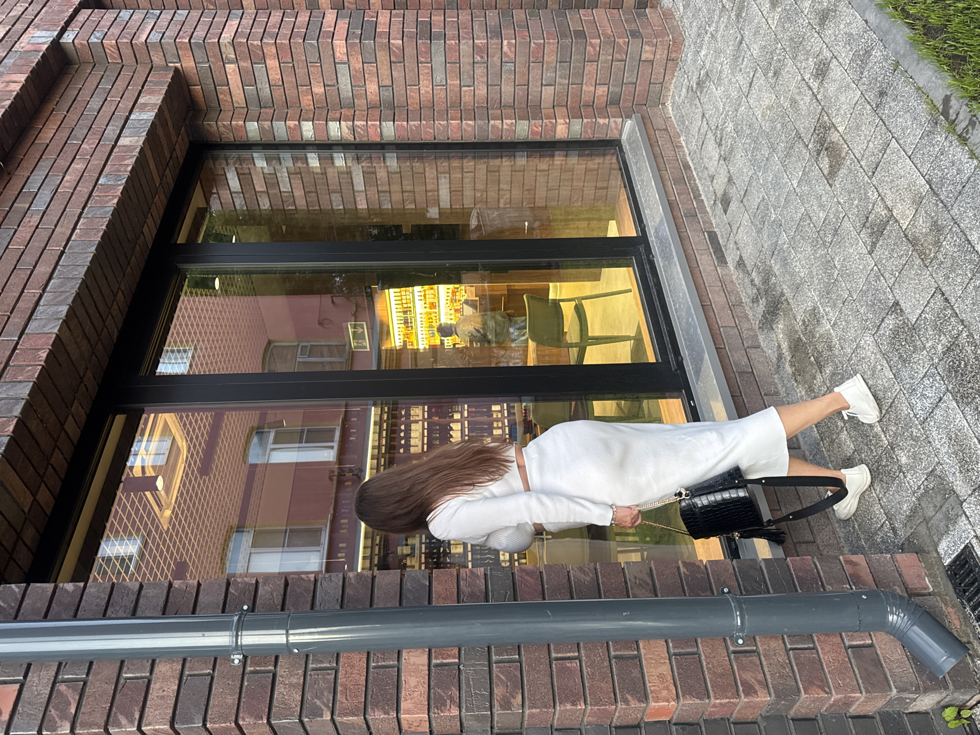 A woman in a white ribbed crop top and skirt is walking on a cobblestone path next to a brick building with a large glass window. She is carrying a black textured handbag with a gold chain. Inside the window, shelves are stocked with bottles, and a person is visible behind a counter. The scene appears to be in a commercial area of Nizhny Novgorod, Russia, possibly a wine shop or similar establishment. The lighting suggests it is either early morning or late afternoon.