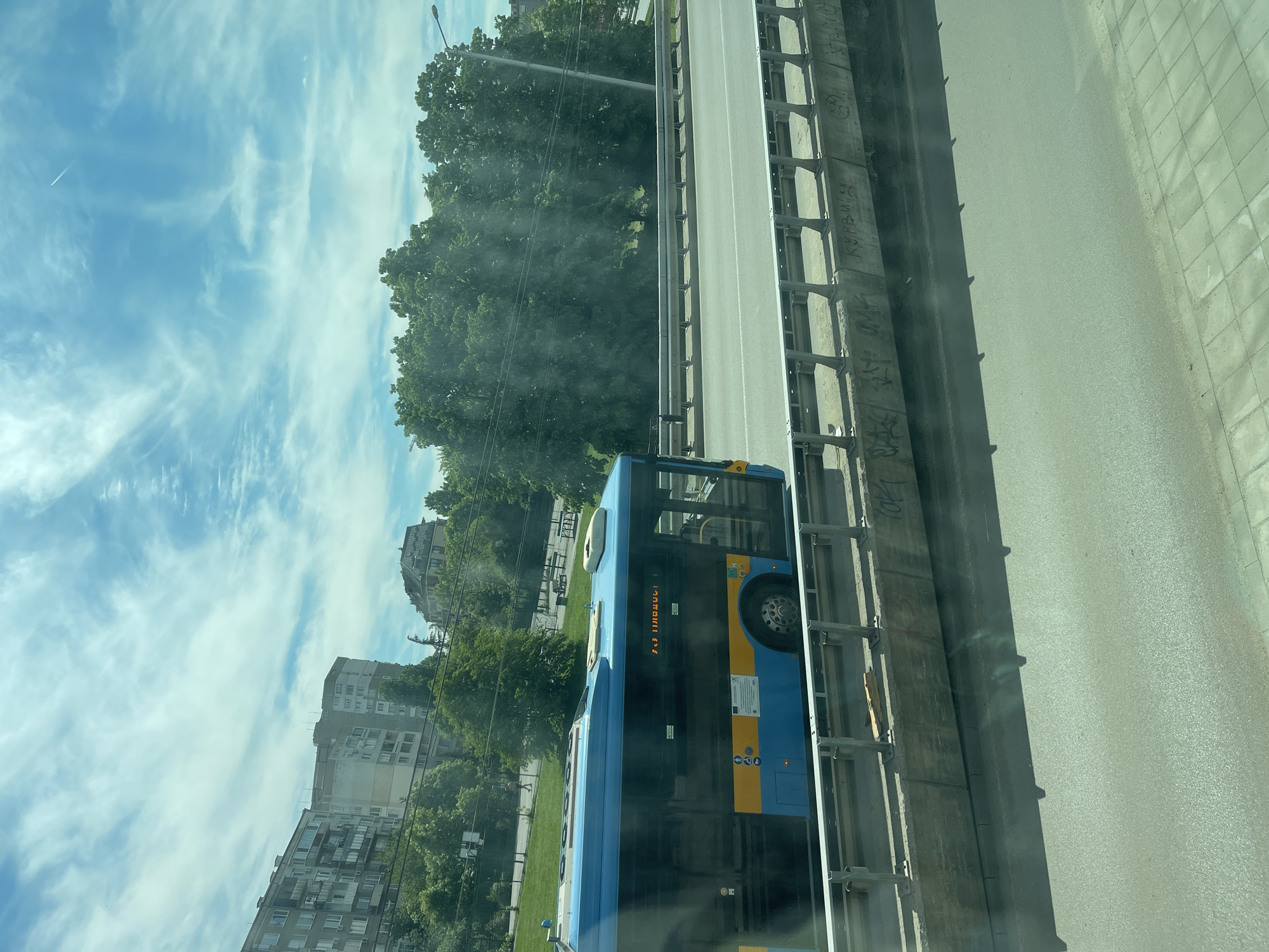 The image shows a blue and yellow public transport bus traveling on a highway in Sofia, Bulgaria. The bus is adjacent to a concrete barrier with graffiti. Beyond the highway, there are green trees and buildings. The sky is blue with scattered clouds. The perspective is from an elevated position.