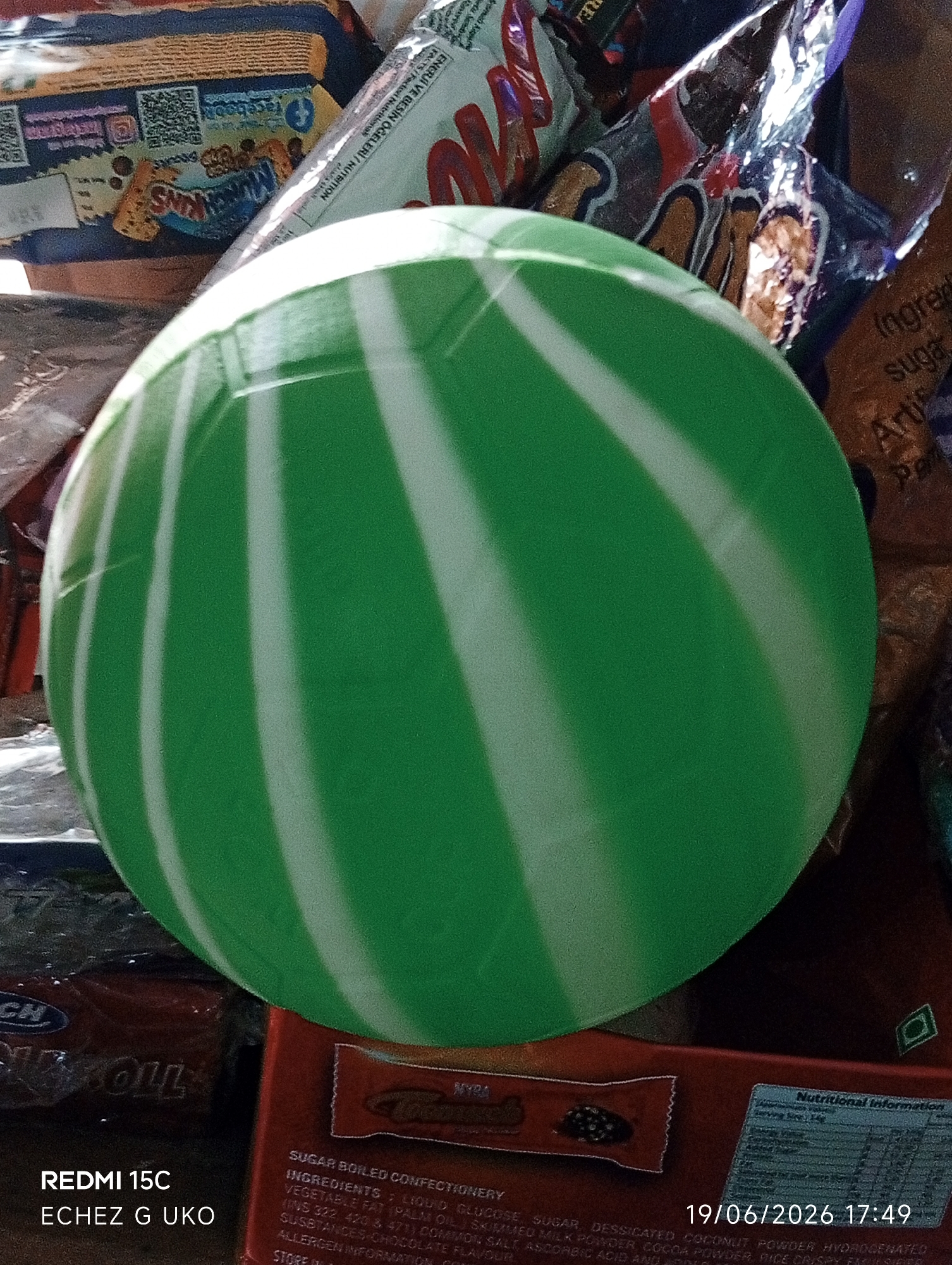 The image shows a green and white striped ball in the foreground, partially obscuring a collection of candy wrappers and packaging. The items are arranged in what appears to be a retail or personal collection setting. In the bottom left corner, text reads "REDMI 15C ECHEZ G UKO". In the bottom right corner, a date and time stamp is visible: "19/06/2026 17:49". The background consists of various colorful wrappers, some with visible text and logos, suggesting a variety of confectionery items. The lighting appears to be natural and somewhat diffused. There is no identifiable human presence or activity within the frame. The scene is static and appears to be a still-life arrangement of objects.