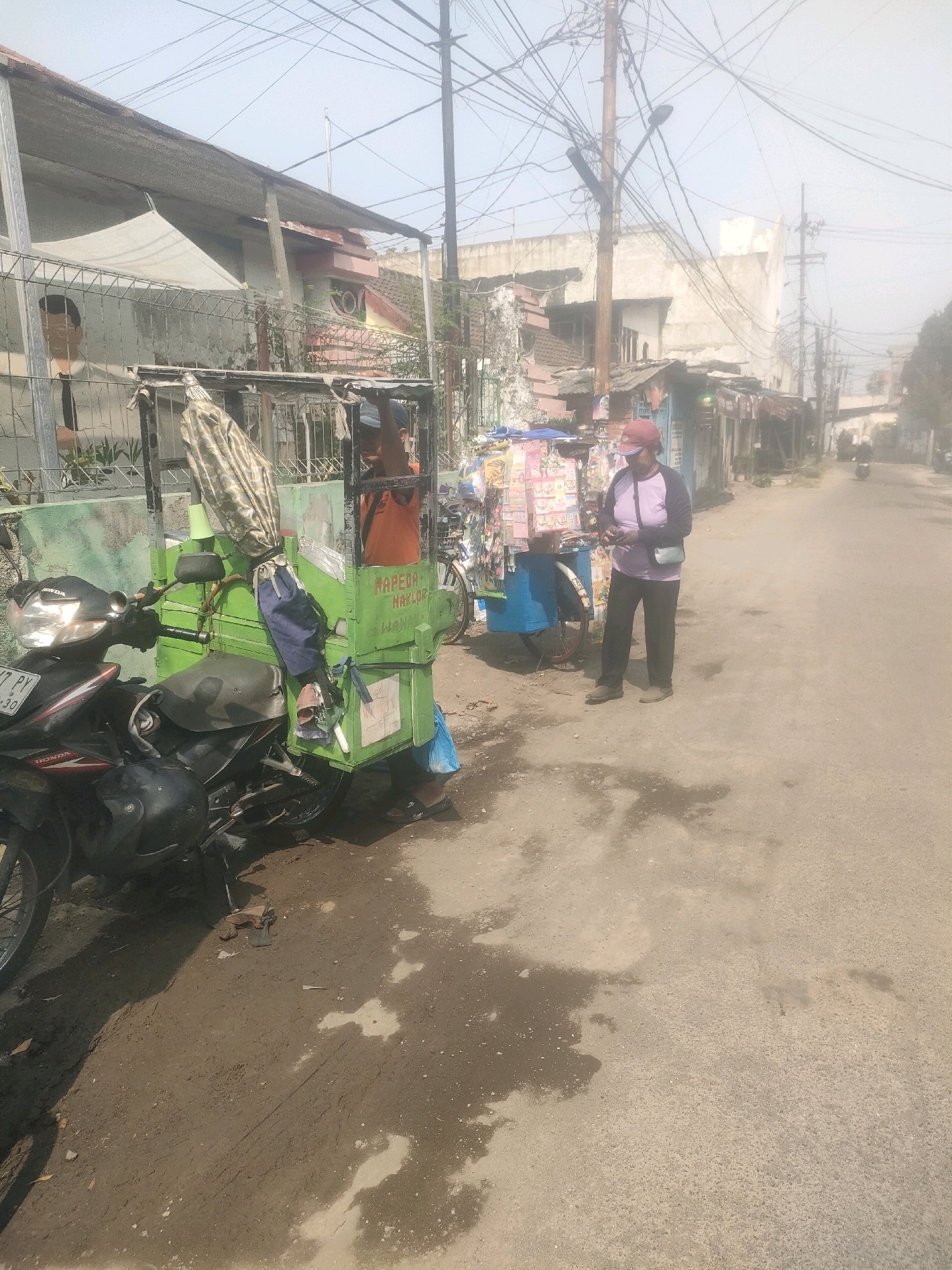 In an urban environment in Surabaya, Indonesia, on a dusty, sunlit street, a person wearing a red cap, a purple long-sleeved shirt, and dark pants stands next to a mobile cart. The cart, a repurposed motorcycle with a green cargo box attached, is loaded with various items, including packaged goods and what appears to be umbrellas. The cart has the text "MAPEDA MAKMUR" and "HANNI" printed on it. To the left of the cart, a motorcycle is parked. In the background, there are several buildings, some with corrugated metal roofs and exposed wiring, suggesting a densely populated area. The street is worn and shows signs of wear and tear, with patches of dry dirt and some wet spots. The overall atmosphere is of everyday street life in a bustling Indonesian city.