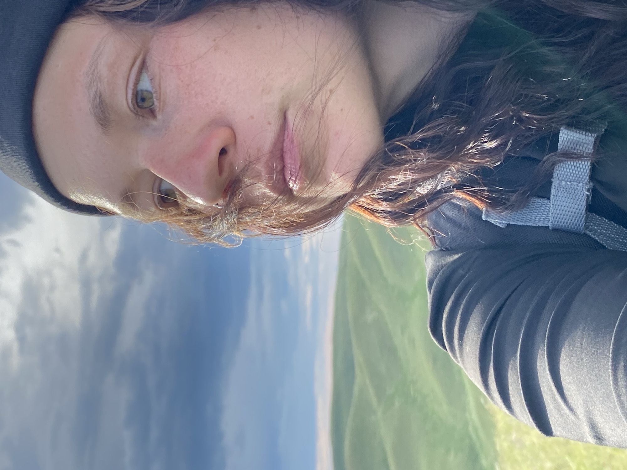 A close-up, slightly angled shot shows a young woman wearing a dark beanie and a dark shirt with a grey strap visible across her chest. Her face, partially obscured by strands of brown hair, is turned to the left, looking towards a hilly, green landscape under a cloudy sky. Her skin has a light complexion with freckles visible on her cheeks and nose. The background features rolling green hills and a sky with dramatic clouds. The overall scene appears to be outdoors in a natural setting. The location is Verkhneozernoe, Russia.