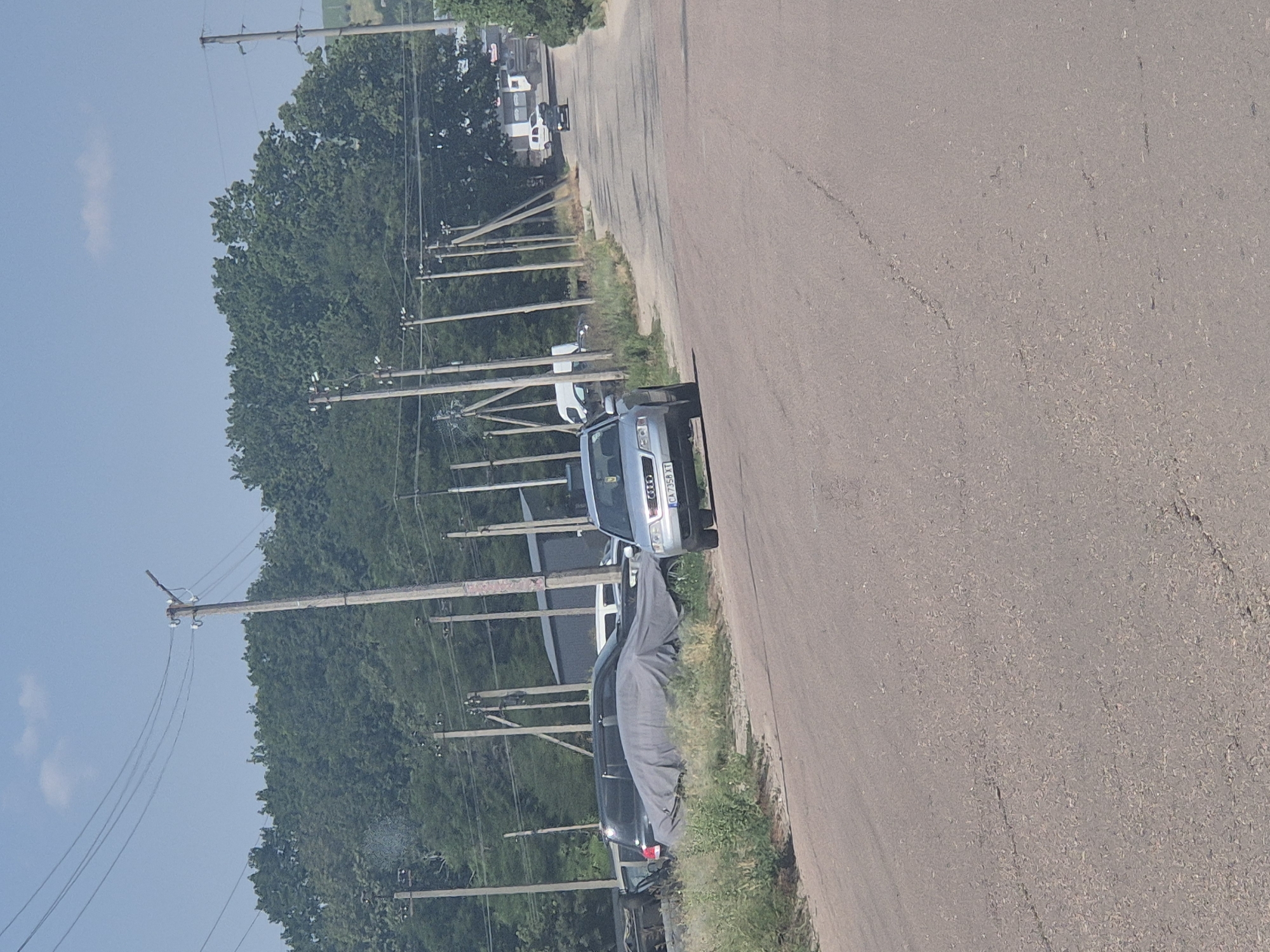 The image depicts a road in Pidhaitsi, Ukraine, with a line of utility poles and trees on the left side. A silver Audi sedan with a license plate reading "CA 7356 XT" is parked on the right side of the road, facing away from the camera. Several other vehicles are visible further down the road, including a white van and a dark-colored car. The road surface is asphalt with visible cracks and patches. The scene appears to be a rural or suburban area on a sunny day with a blue sky and scattered clouds.