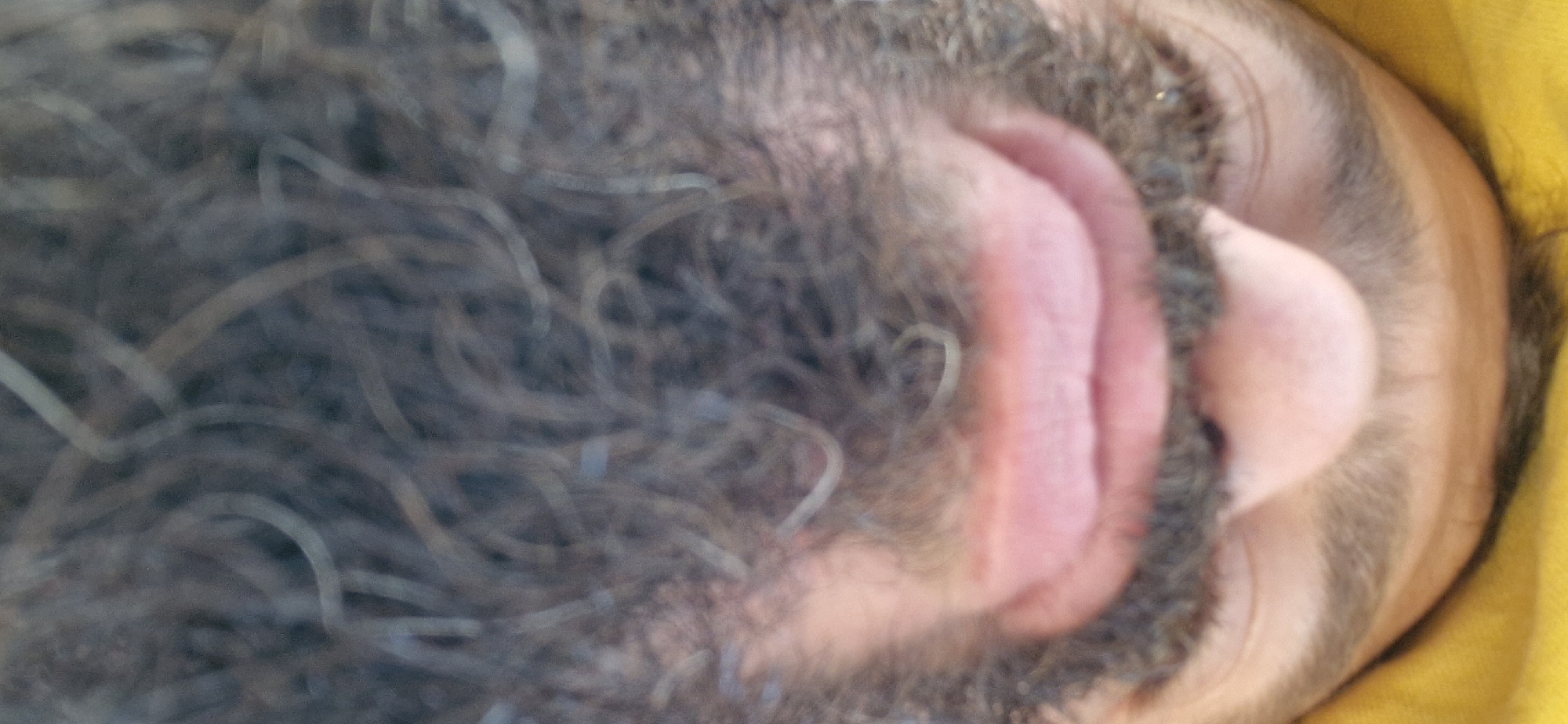 The image is an extreme close-up, upside-down, and low-angle shot of a person's face, specifically focusing on their nose, mouth, and beard, with a yellow fabric visible at the top. The lighting is diffuse, and the image is slightly out of focus, especially in the lower portion. There are no discernible features of Wattenwil, Switzerland, or any urban environment. The scene is intimate and personal, with the subject's skin texture and facial hair clearly visible.