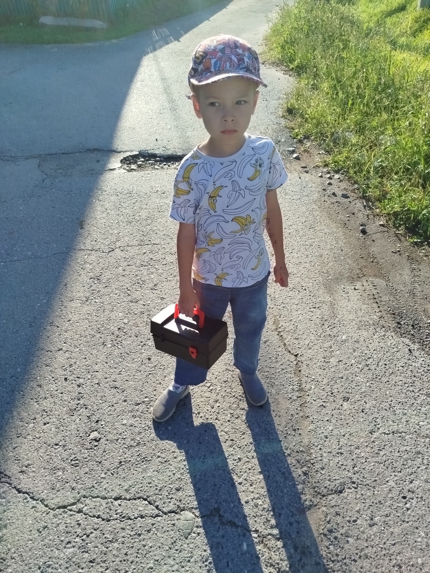 A young boy, approximately 5-7 years old, stands on a cracked asphalt road in Zavodoukovsk, Russia. He is wearing a baseball cap, a white t-shirt with a banana print, blue jeans, and grey slip-on shoes. The boy is holding a black toolbox with red handles. Sunlight casts a long shadow to his left. The road is bordered by a grassy area on the right and a green fence on the left, suggesting a rural or suburban setting. The scene appears to be captured in the late afternoon given the angle of the sun and the length of the shadows.