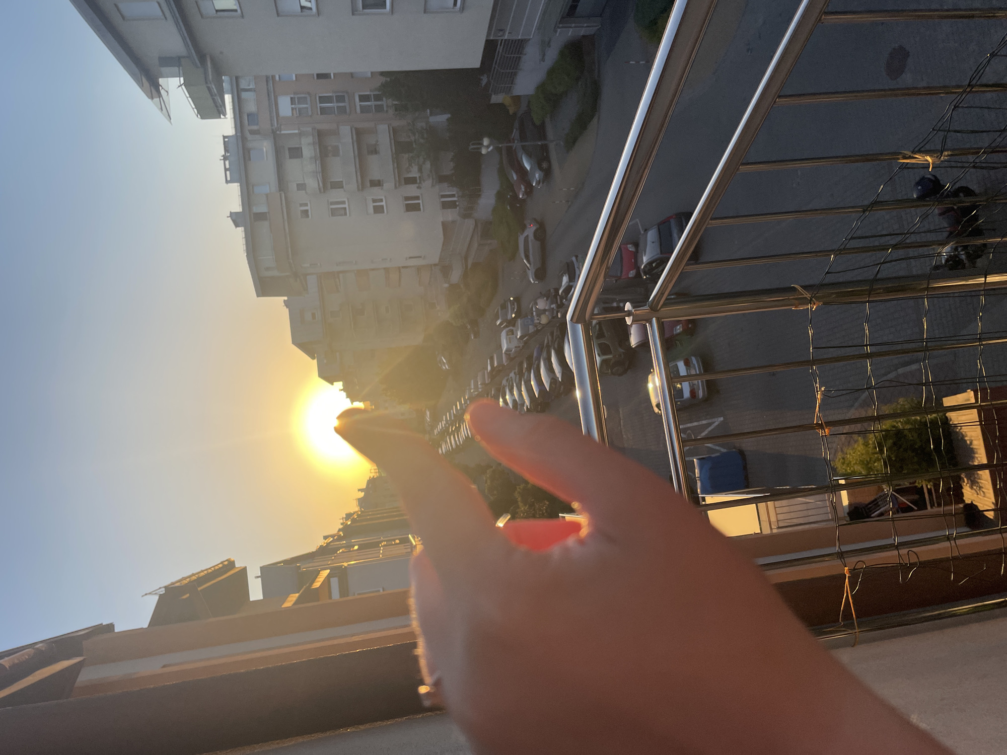 Here is a forensic description of the image:
The image shows a cityscape at sunset from a high vantage point, possibly a balcony. The sun is low on the horizon, casting a strong orange glow. There are multi-story buildings visible in the background. A hand is in the foreground, appearing to reach for the sun. A metal railing is also visible, indicating the viewpoint is likely a balcony. Cars are parked along the street below. There are also many trees. The image appears to be taken in a densely populated urban area.