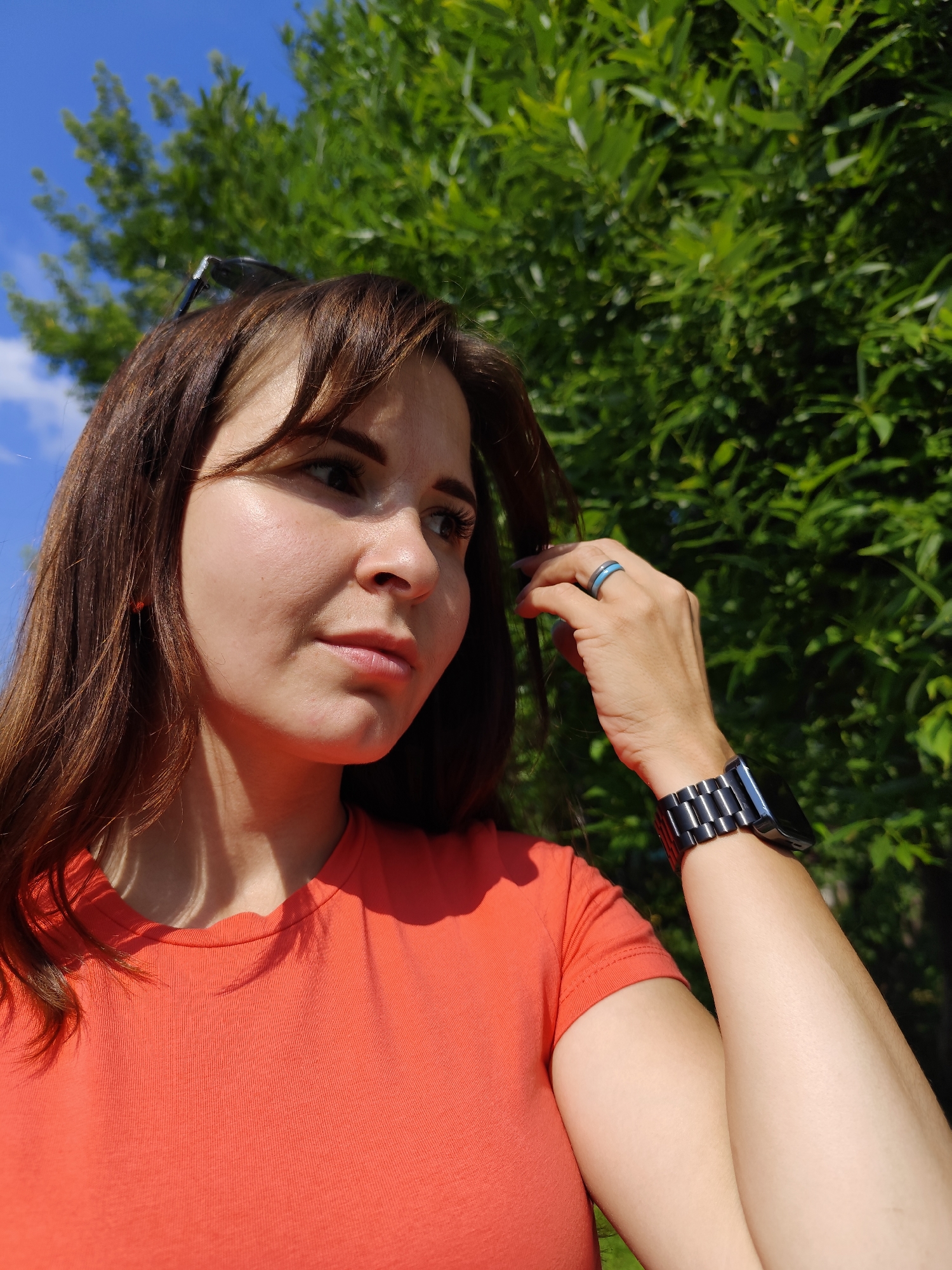 A medium shot captures a woman with brown hair and light skin, wearing an orange t-shirt and a smartwatch. She is outdoors, with a bright blue sky and lush green foliage in the background. Her left hand is near her face, touching her hair. The woman is looking to her right, her expression neutral. The lighting suggests it is daytime, with sunlight casting shadows on her face and t-shirt. The location is identified as Foki, Russia.