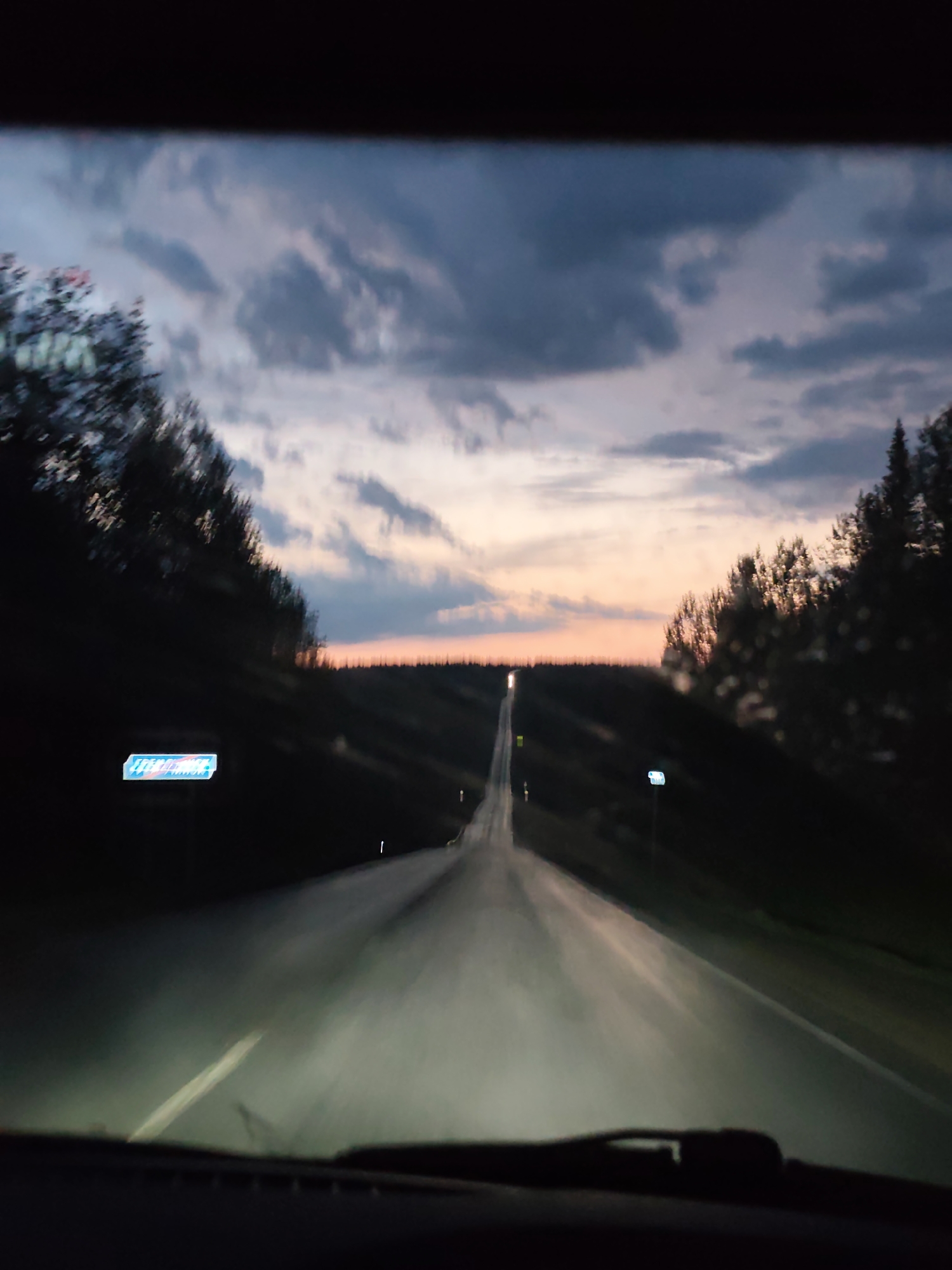 The image depicts a road in Gremyachinsk, Russia, at dusk or dawn, taken from the interior of a vehicle. The road stretches into the distance, flanked by trees on both sides. The sky is a mix of dark and light clouds, with a horizon suggesting the beginning or end of the day. The road surface appears wet, reflecting the headlights of the vehicle. A road sign is visible on the left side of the road, while another is on the right. The windshield wipers are visible at the bottom of the frame. The overall scene is dark, illuminated mainly by the vehicle's headlights and the twilight sky.
