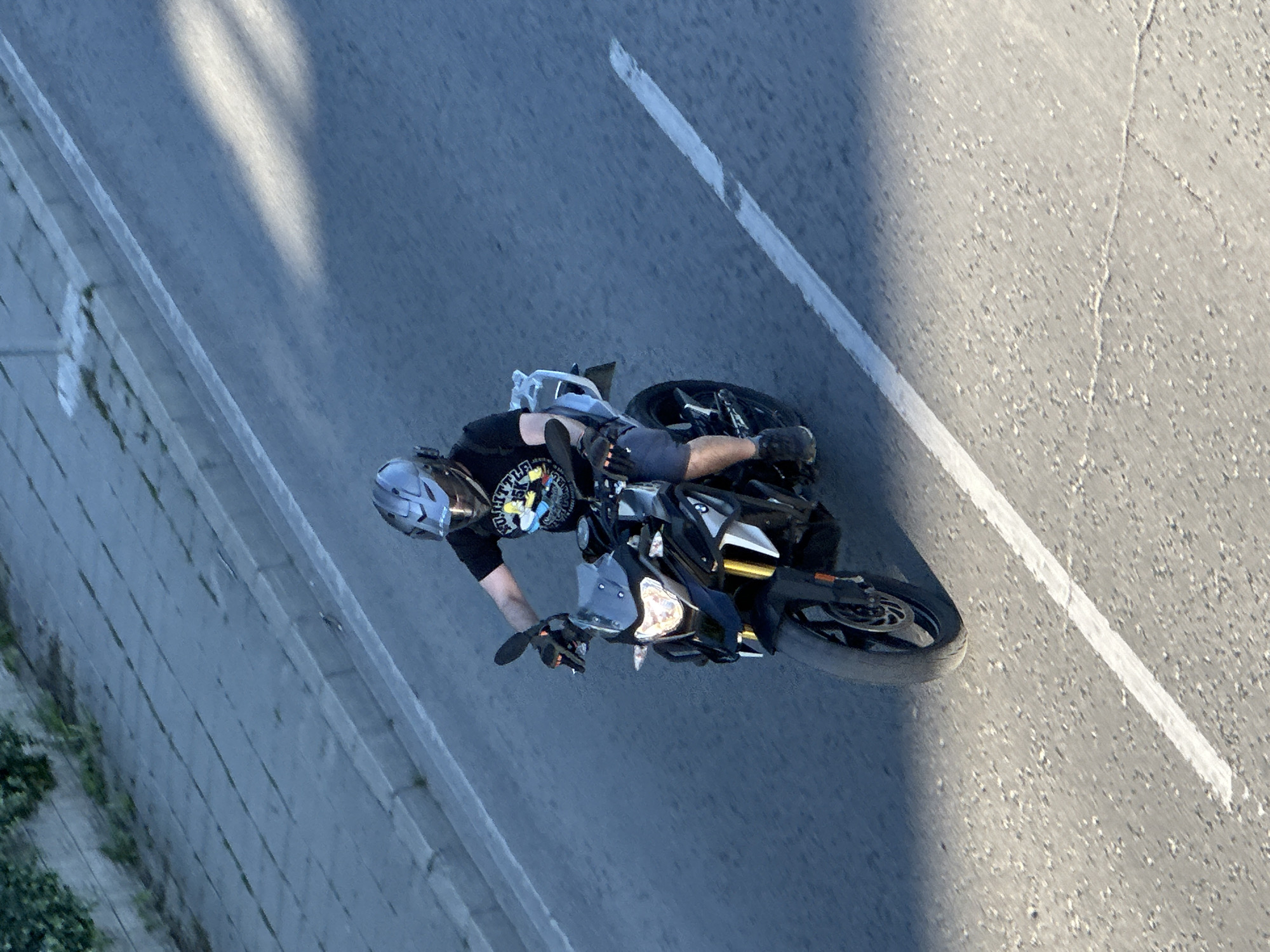 An aerial shot shows a motorcyclist riding a black motorcycle on an asphalt road. The rider wears a helmet, black t-shirt, grey shorts, and gloves. A white line is painted on the road next to the motorcycle. The road is situated in the city of Veliko Tarnovo, Bulgaria.